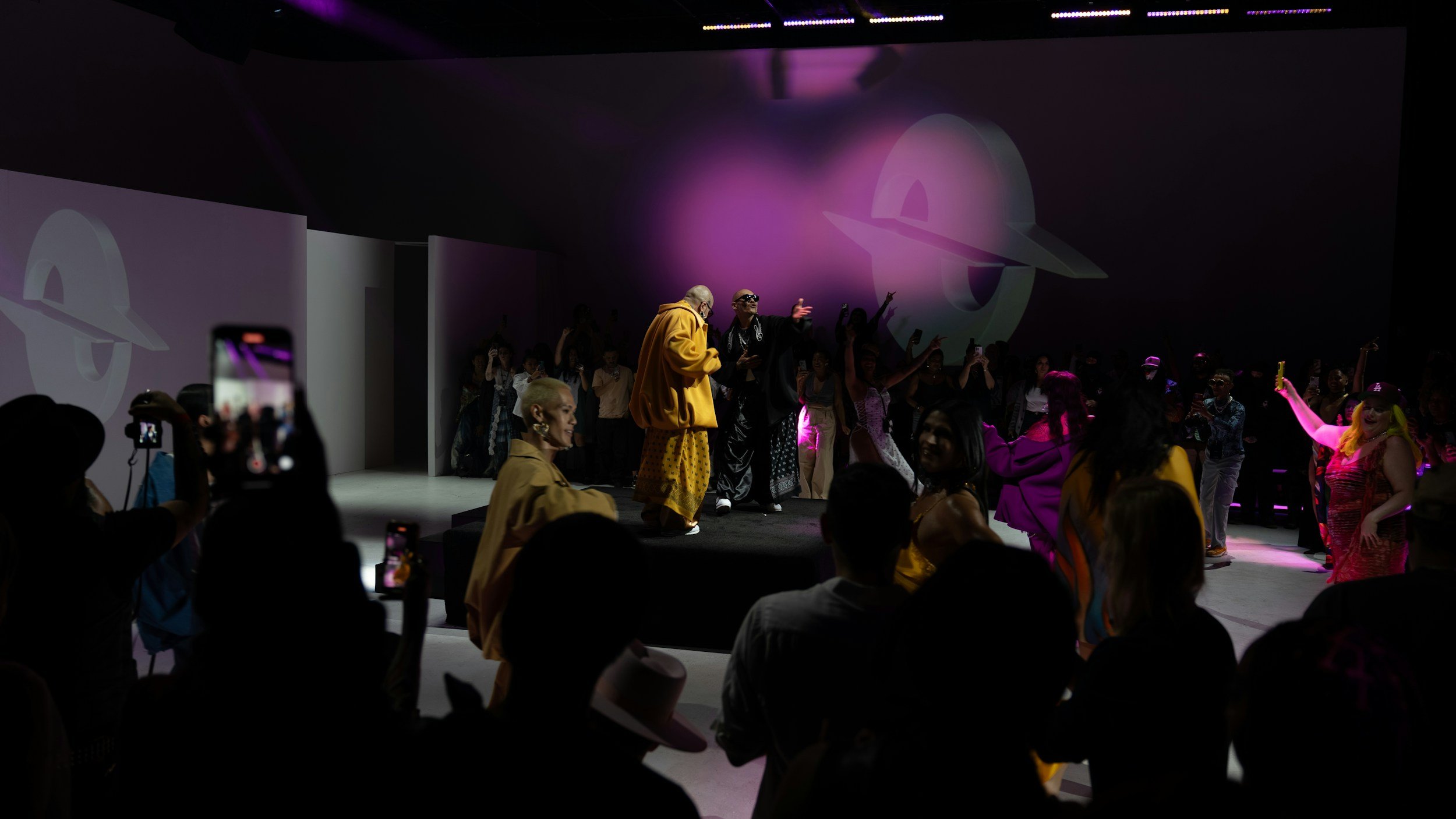 People dancing and posing at a fashion show or event on a stage with a purple logo projection in the background, surrounded by an audience taking photos.