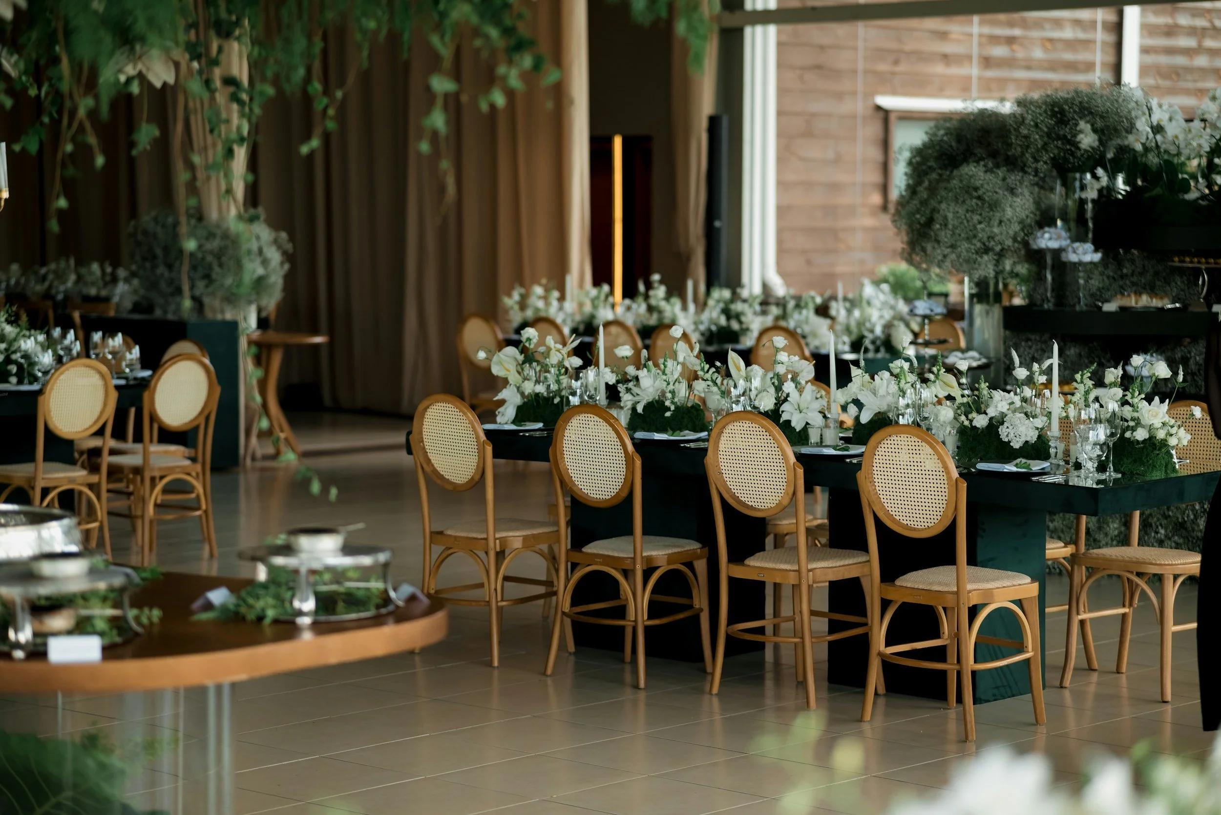 Elegant indoor event setup with round tables decorated with white floral arrangements and tall candles, surrounded by wooden bistro chairs. The background features a wooden wall, greenery, and a curtain, creating a warm and inviting atmosphere for a celebration or gathering.