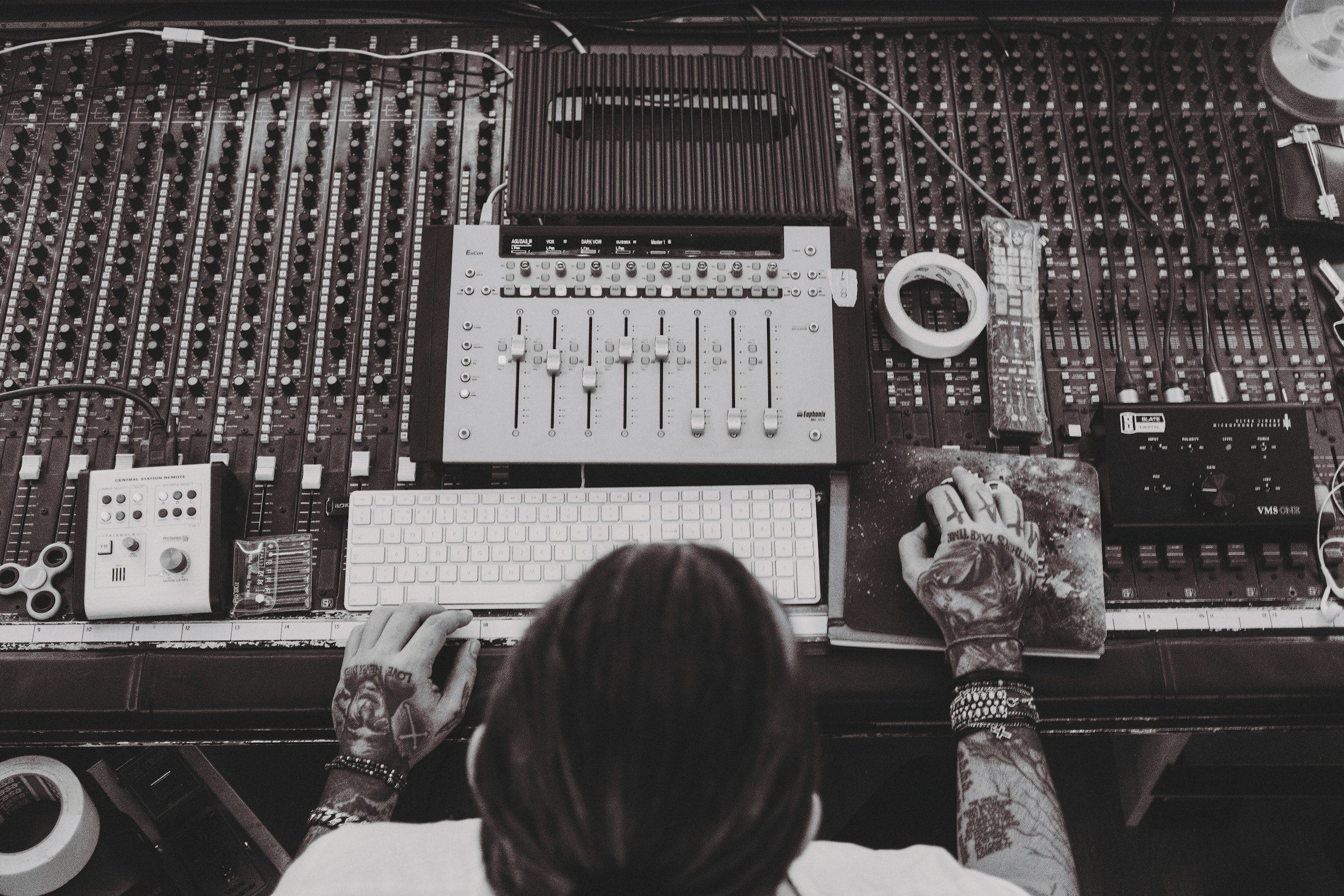 A top-down view of a music production setup with a mixing console, a computer keyboard, and various electronic music equipment. Two tattooed hands are working on the equipment, with one hand near a MIDI controller and the other on the mixing console.