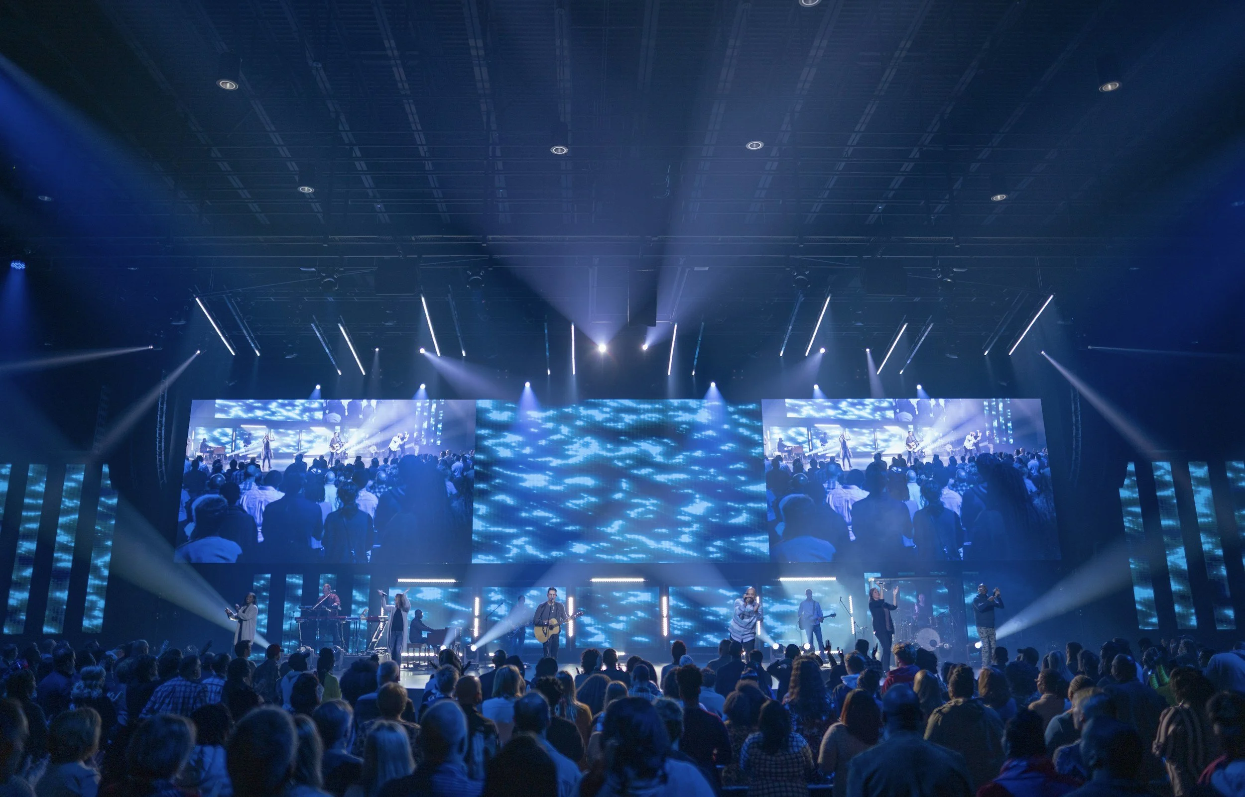 A large indoor concert or event with a stage illuminated by blue and white lights. The audience is standing and watching a live band or musical performance, with multiple musicians visible on stage.