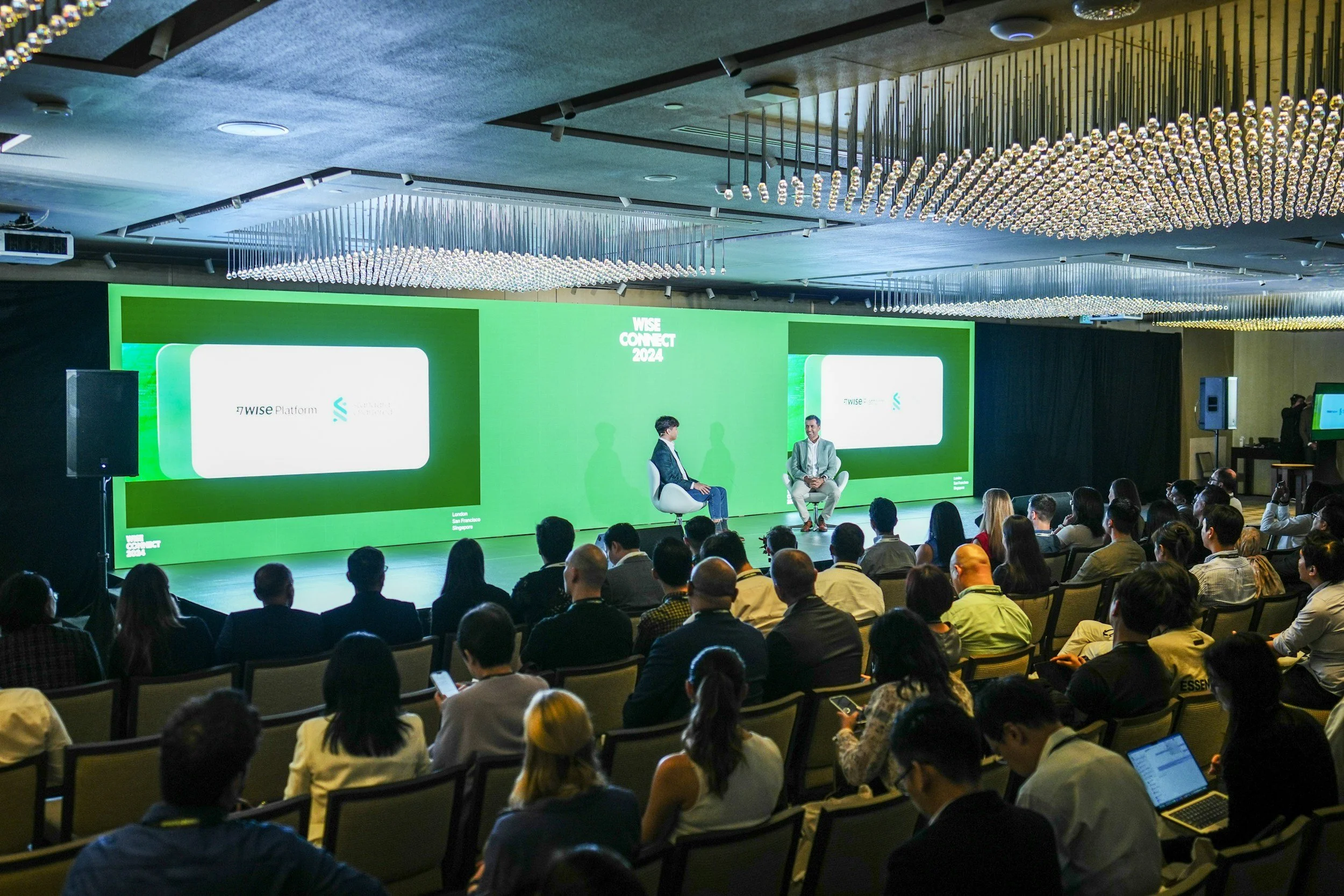 A large conference room filled with seated audience members facing a stage. On stage, two men are sitting and engaging in a discussion. Behind them, there is a large green digital screen displaying the event name 'WISE CONNECT 2024' and the logo of WISE Platform. The ceiling has decorative light fixtures with numerous small lights.