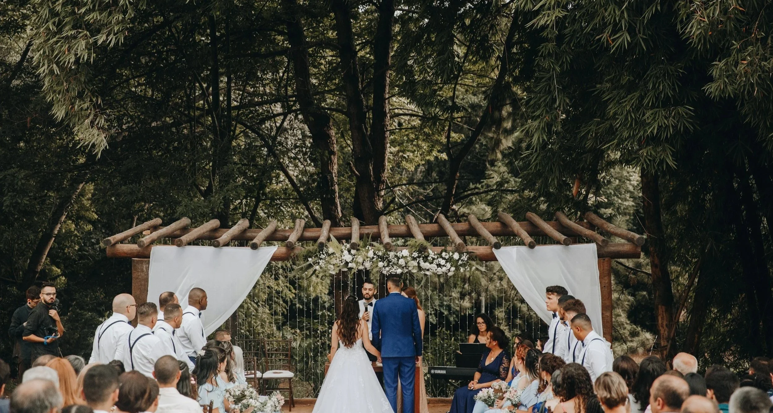 A couple at their wedding ceremony outdoors under a wooden arch decorated with white curtains and flowers, with wedding guests seated around them and tall trees in the background.