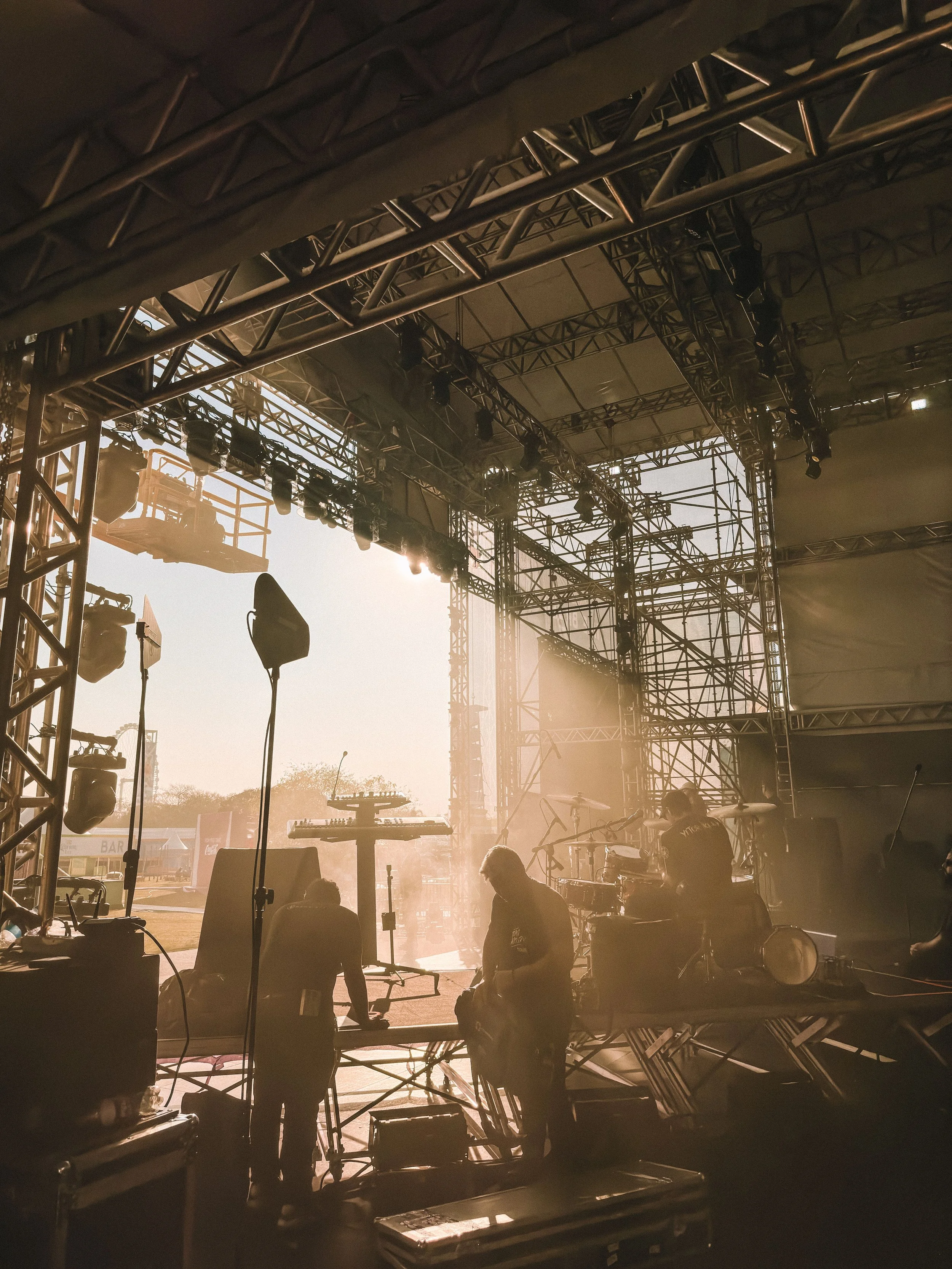 Stage setup with lighting and scaffolding, likely at a concert or festival, with silhouettes of crew working backstage as sunlight filters through.