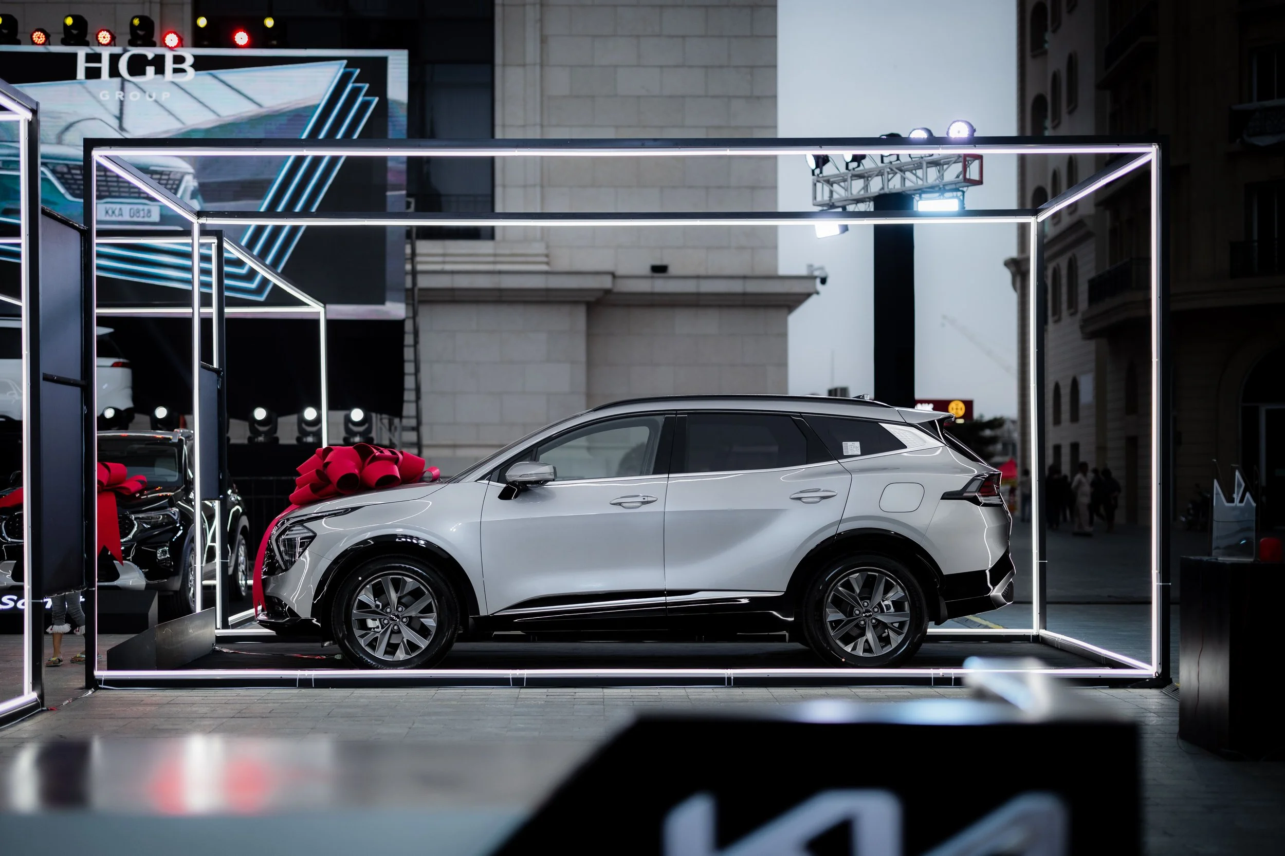 A silver SUV displayed at an outdoor auto show under a illuminated frame with a red ribbon, with several other cars and a large digital screen in the background.