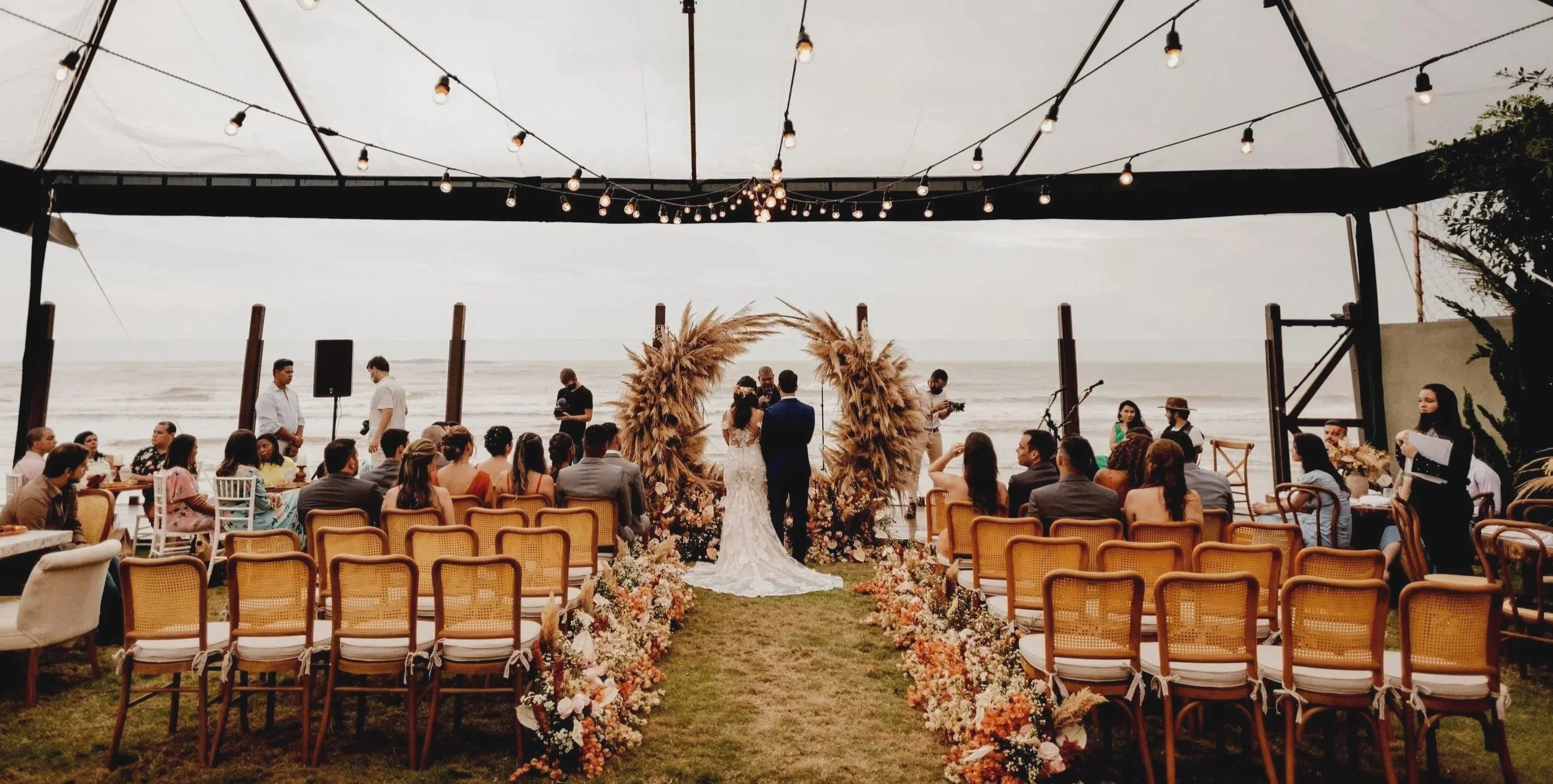 A beachside outdoor wedding ceremony with a decorated arch, guests seated under a canopy with string lights, and the ocean in the background.