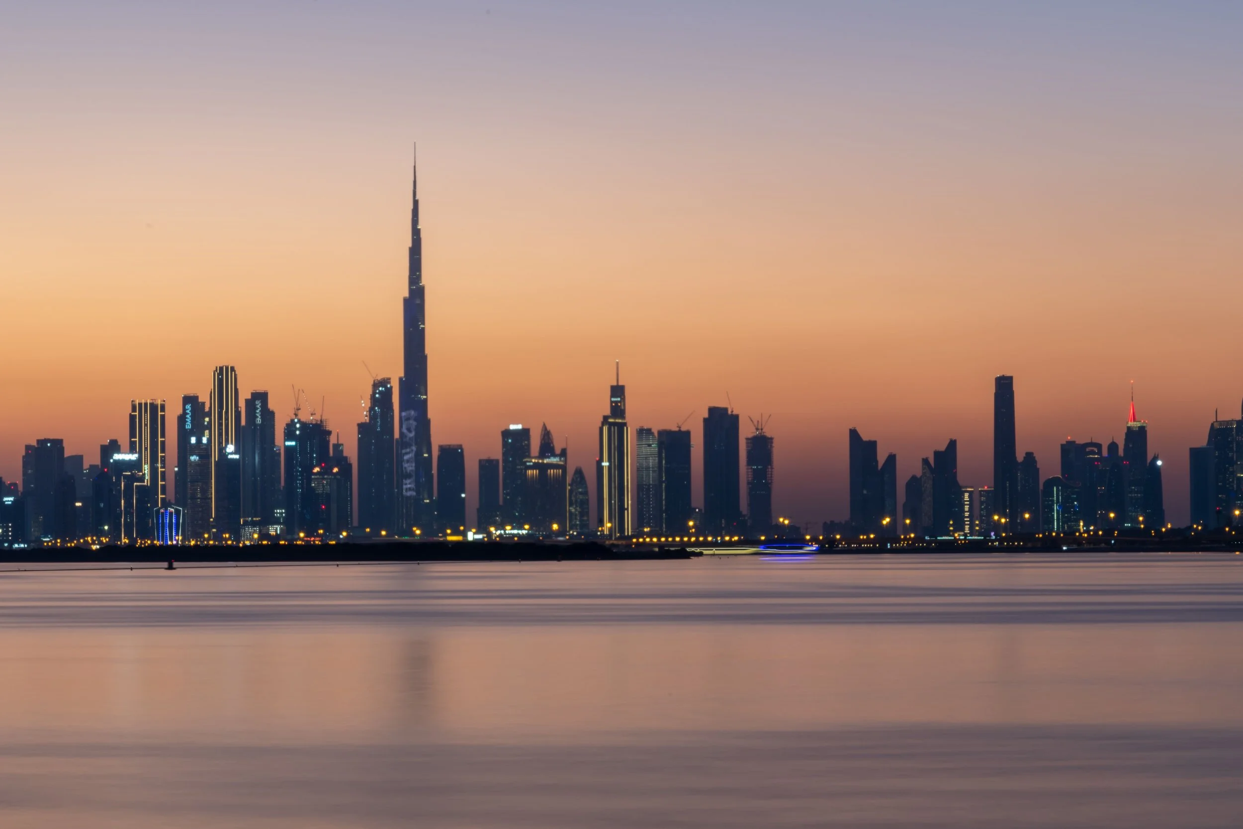 City skyline during sunset with tall buildings, including the Burj Khalifa, reflecting city lights across a body of water.