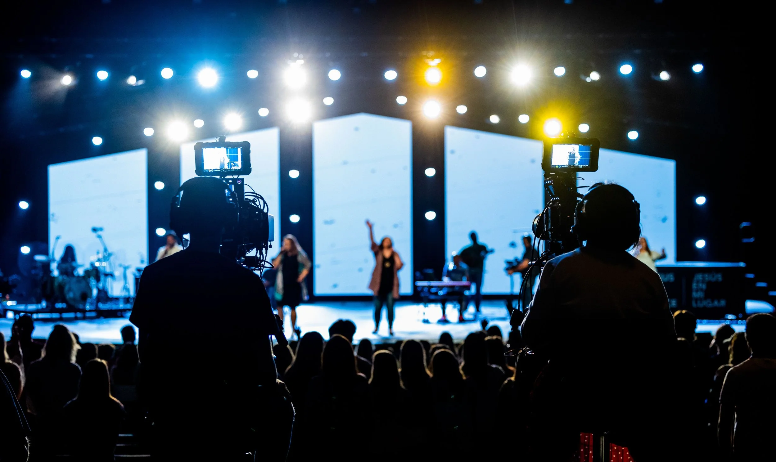 A stage with speakers and a band performing, brightly lit with multiple spotlights, audience silhouettes in the foreground, and camera operators capturing the event.