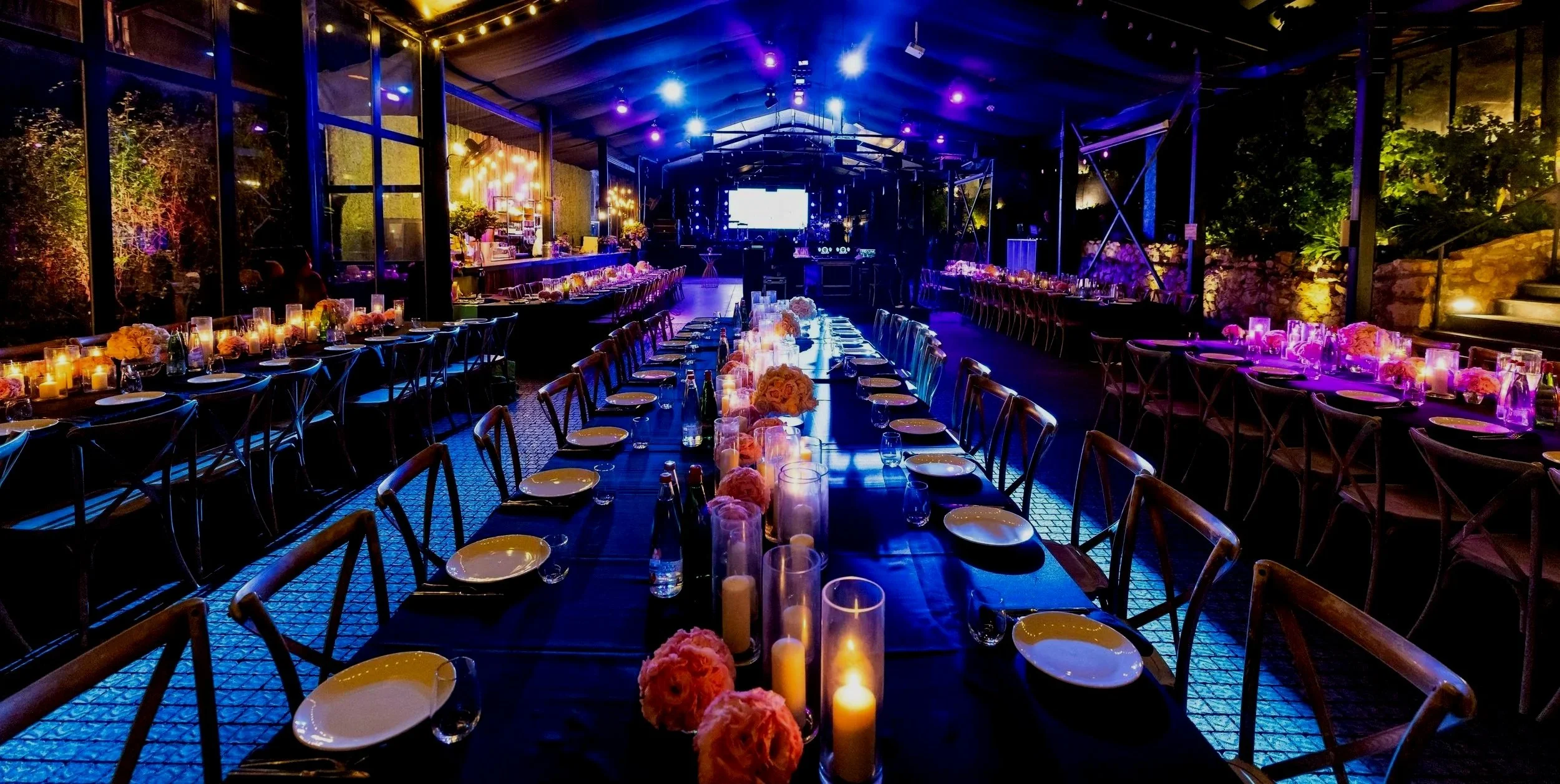 Indoor event space decorated with candles and flowers, set up for a dinner or celebration, with tables, chairs, and purple lighting.