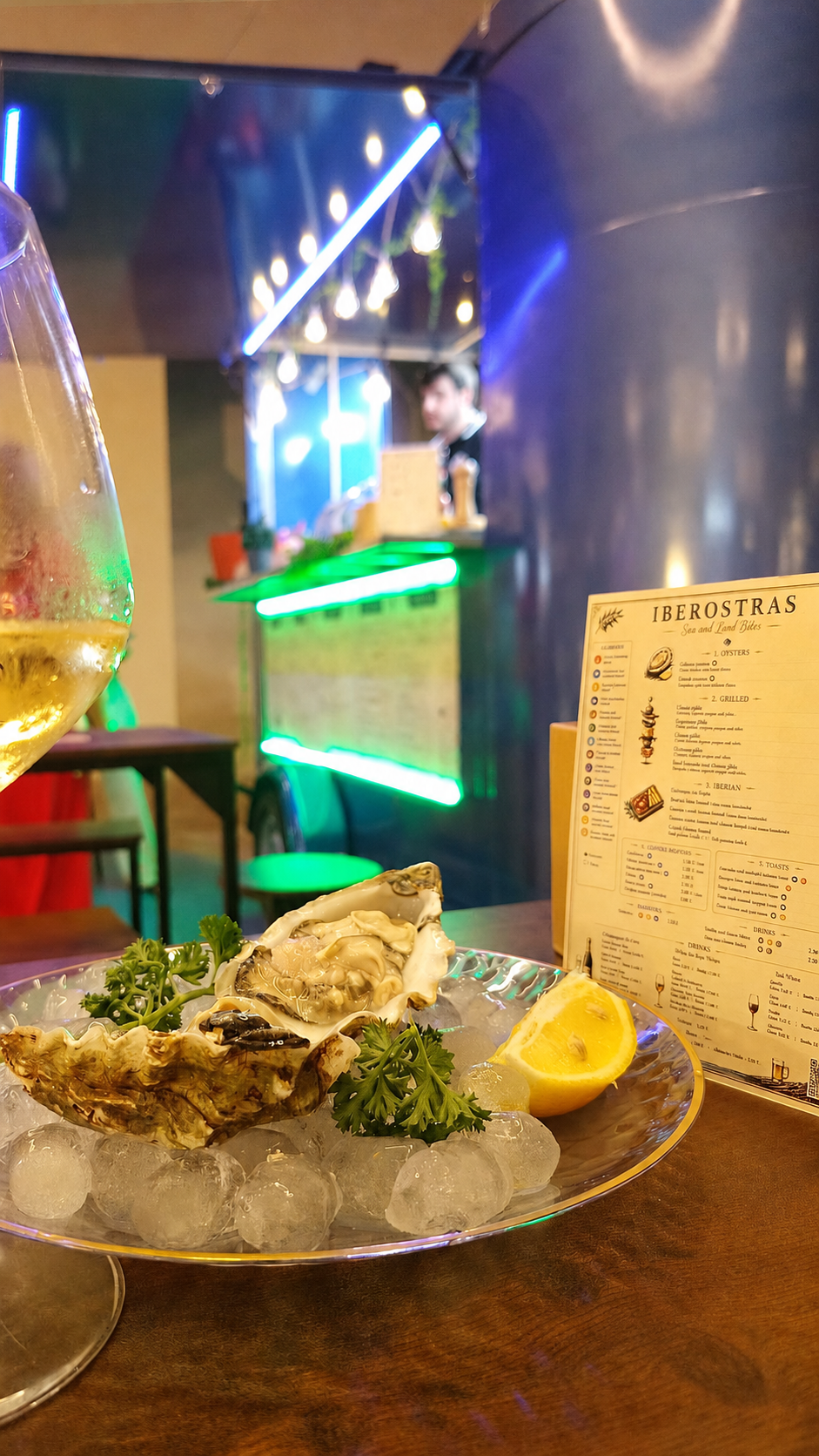 Mariscos en hielo con limón y perejil, una copa de vino y un menú en un restaurante con iluminación colorida y ambiente moderno.