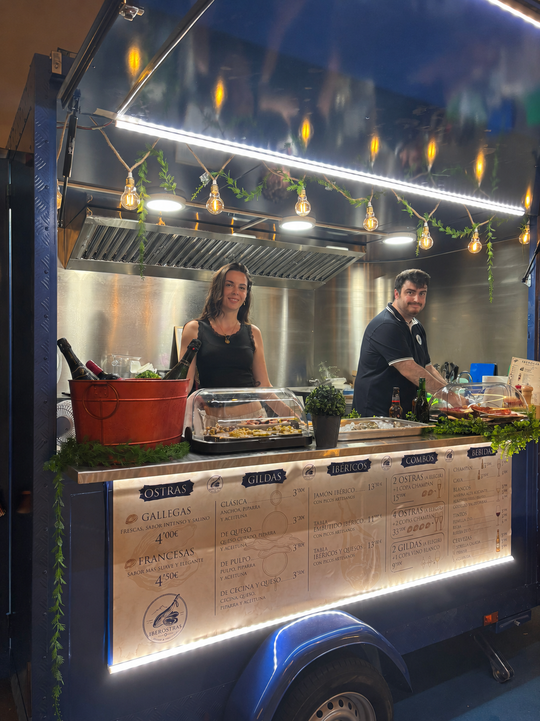 Vendedor de mariscos en una taquería móvil decorada con luces en el techo y plantas verdes, con un cartel de menú que ofrece diferentes tipos de mariscos y combinaciones.