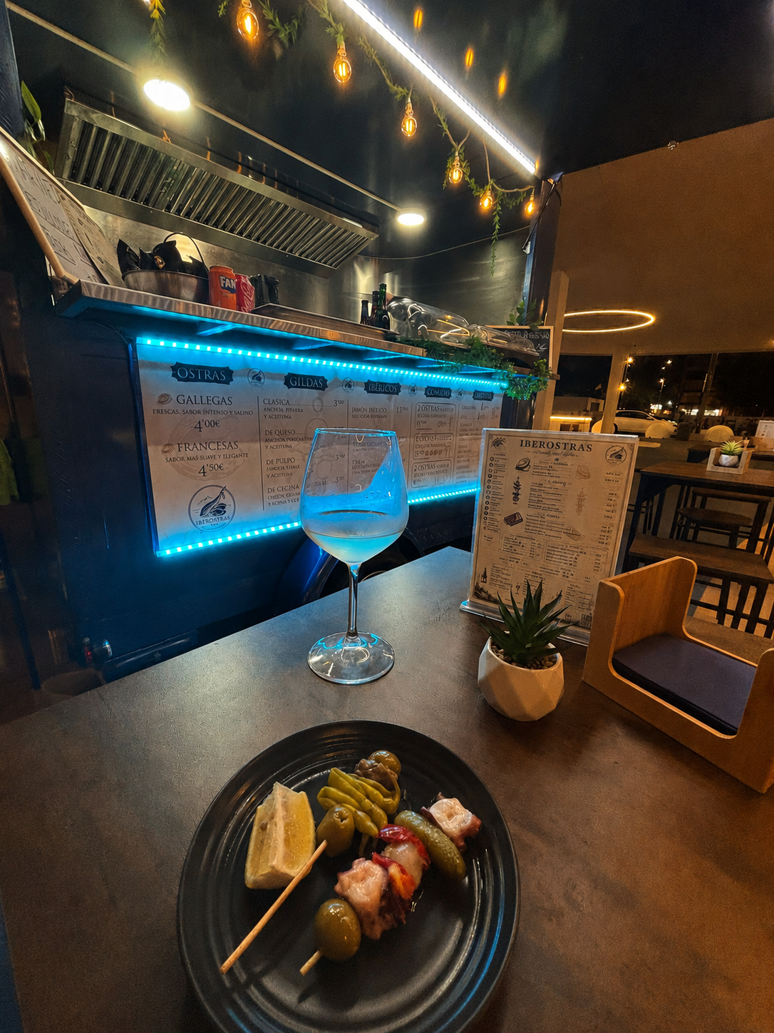 Mesa con un plato de pinchos, una copa de vino y un menú en un restaurante con decoración moderna y luces cálidas.