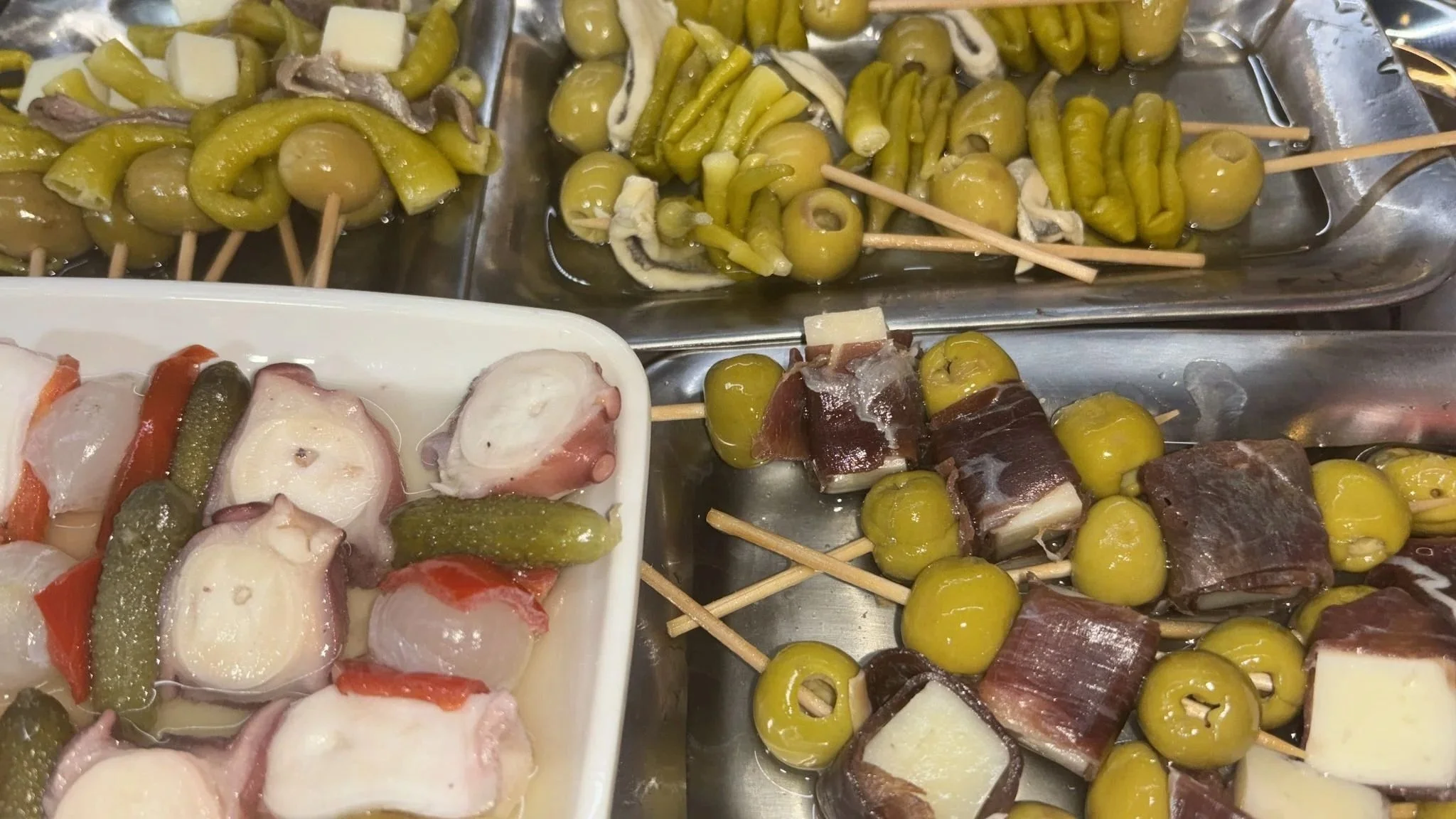 Antojitos de comida tradicional, con varios tipos de aceitunas, pepinos, jamón, queso y otros ingredientes en palitos de madera, en diferentes bandejas.
