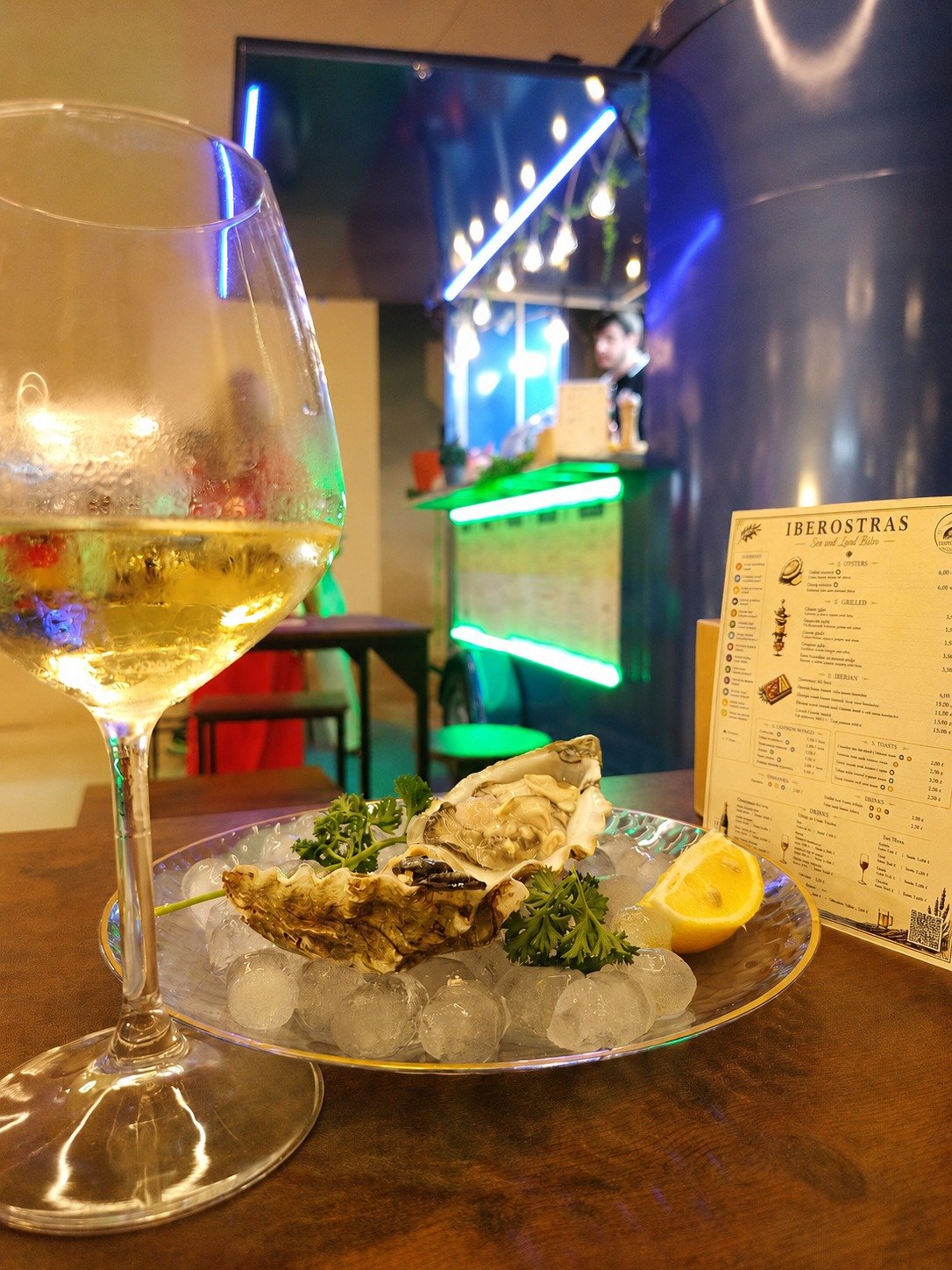 Vasija con vino blanco, plato con ostiones, rodaja de limón y perejil en hielo, fondo de un bar con luces de neón verdes y un menú volado a la derecha.
