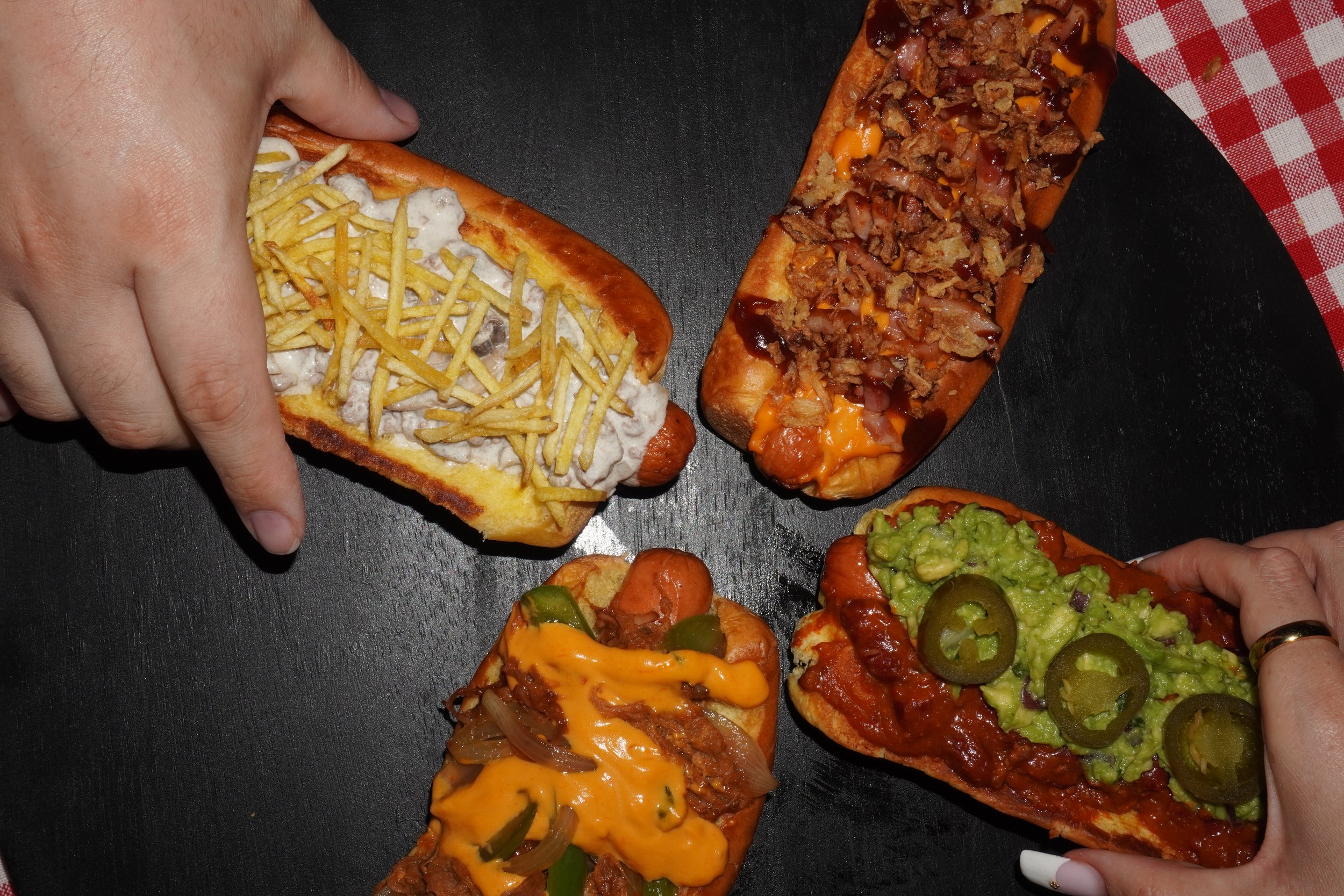 Varios perritos caliente con diferentes ingredientes como carne, verduras, crema y papas, y una mano sujetando una porción de pizza, sobre una superficie de color negro con un mantel a cuadros rojo y blanco.
