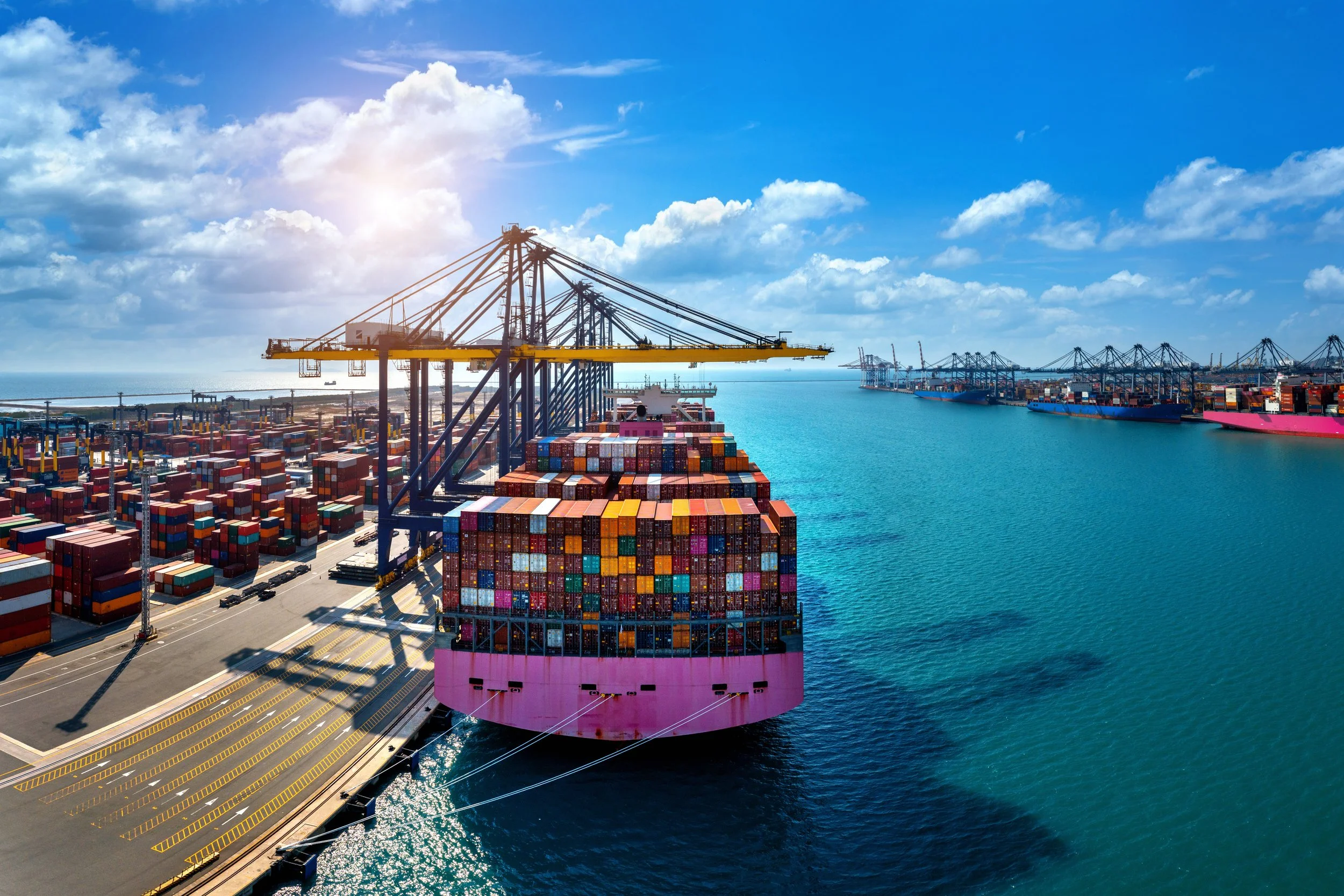 A large cargo ship carrying stacked containers is docked at a port with multiple crane vessels for loading and unloading. The port is next to a body of water under a partly cloudy sky with the sun shining.