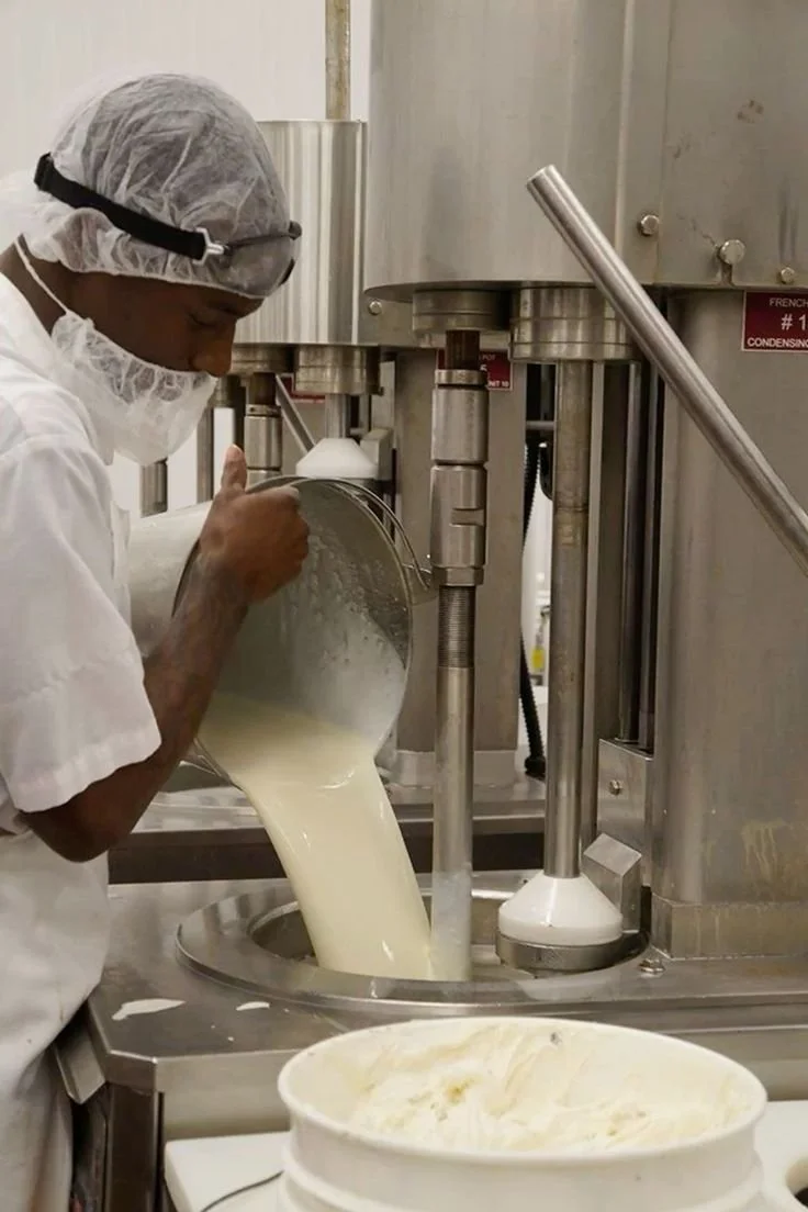 A person in protective gear pouring a liquid mixture into a large industrial machine, likely in a food processing or manufacturing setting.