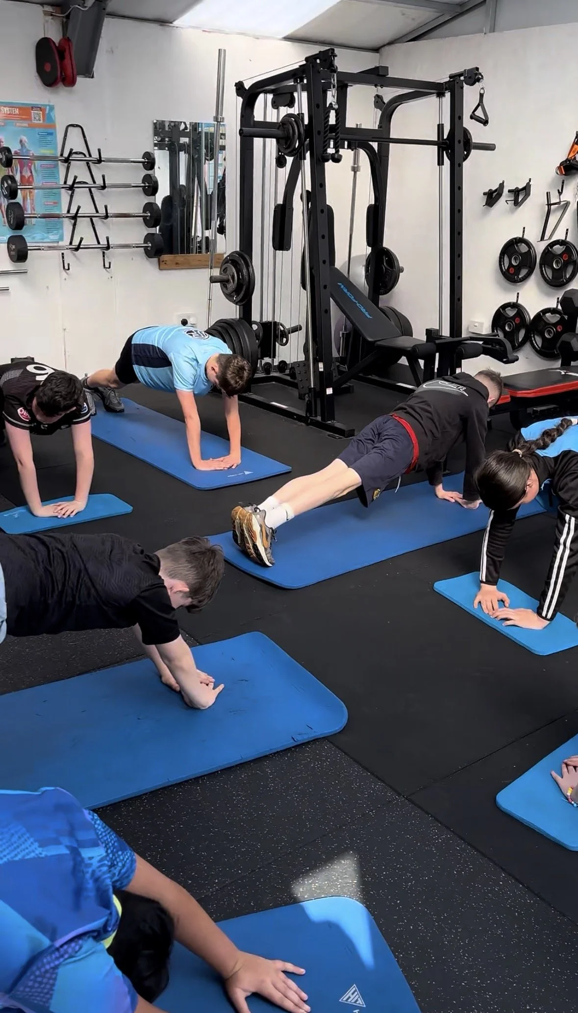 A group of children performing planks on blue exercise mats in a gym with weightlifting equipment in the background.