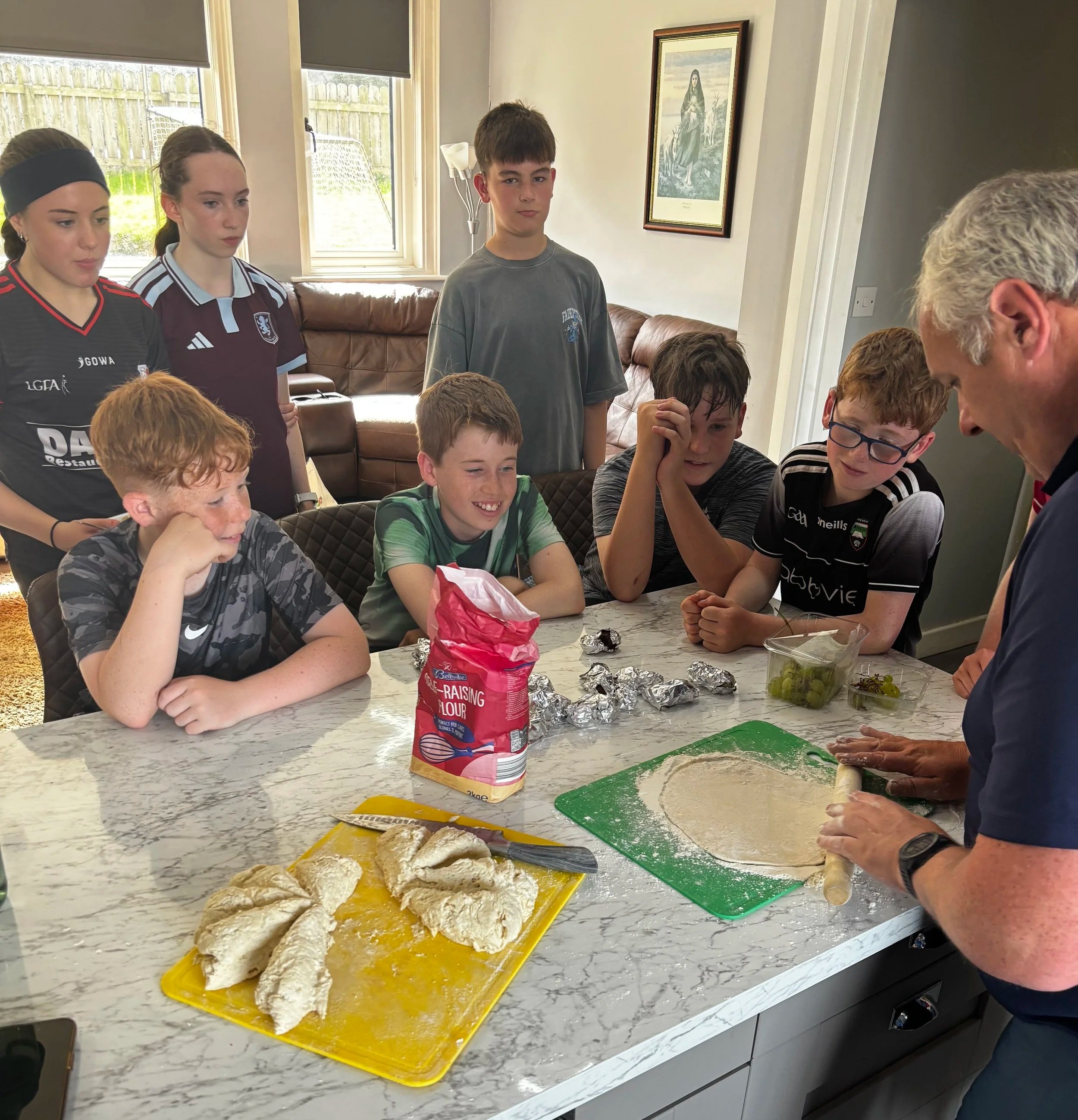 A group of children gathered around a table with food prep ingredients, including a yellow tray with dough, a bag of flour, and wrapped items, in a bright indoor setting with large windows and a painting on the wall.