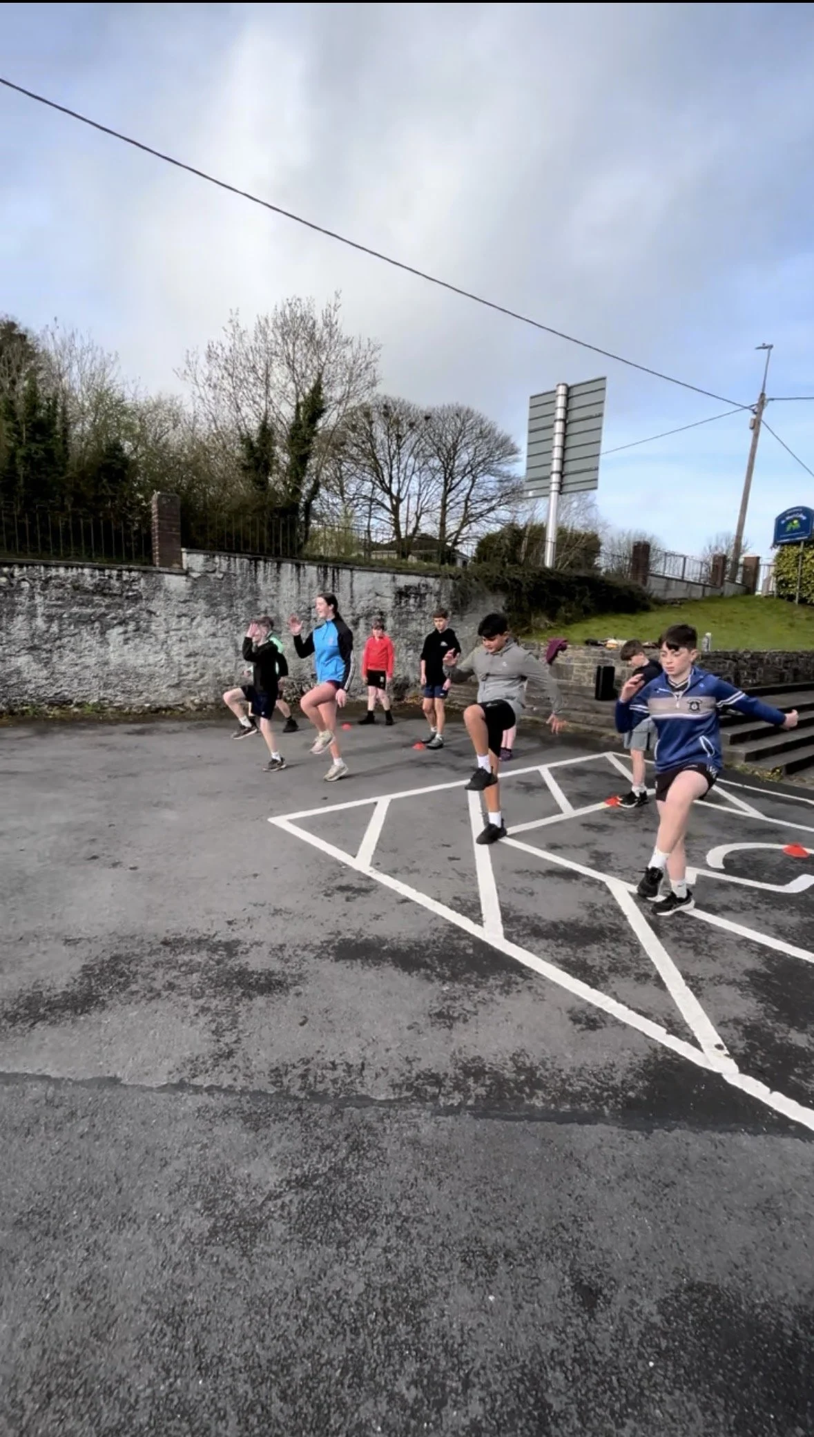 A group of children and teenagers exercising outdoors on a parking lot area marked with painted lines and orange cones.