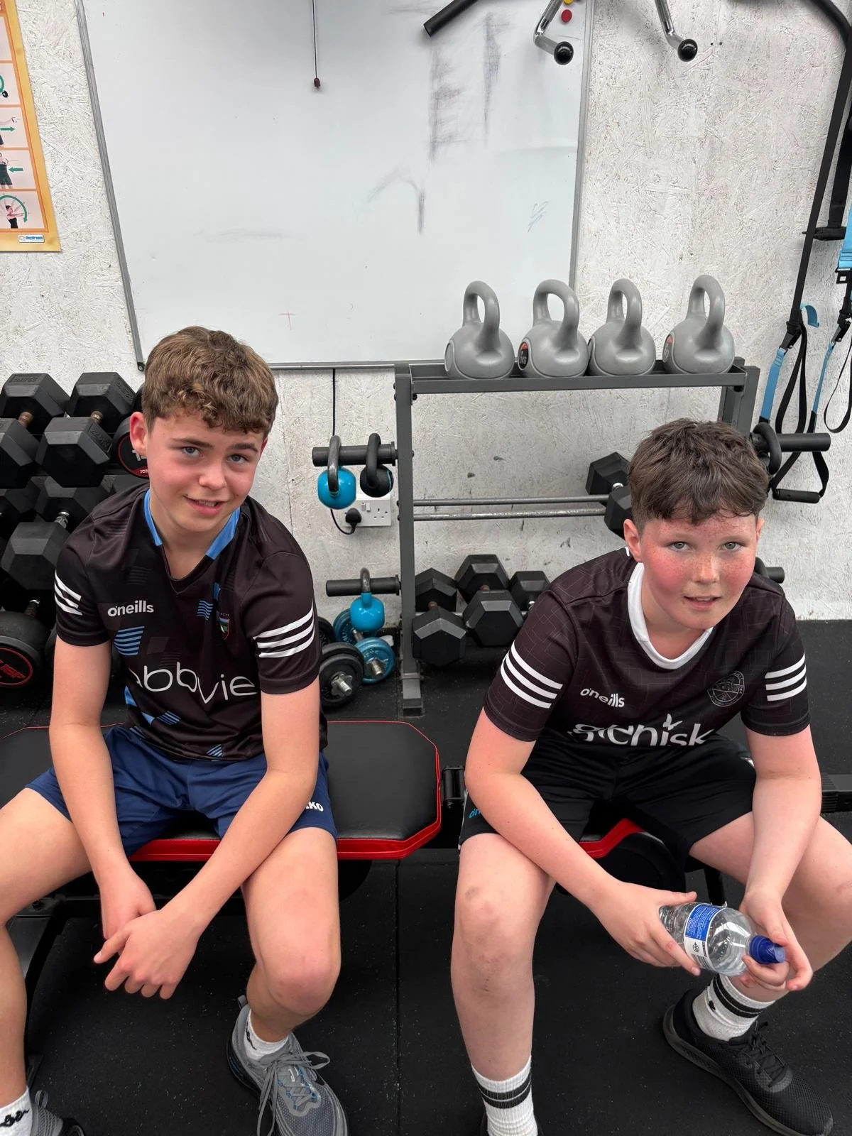 Two young boys in black athletic shirts sitting on workout benches in a gym, surrounded by weights and fitness equipment, with kettlebells on a rack and a whiteboard behind them.