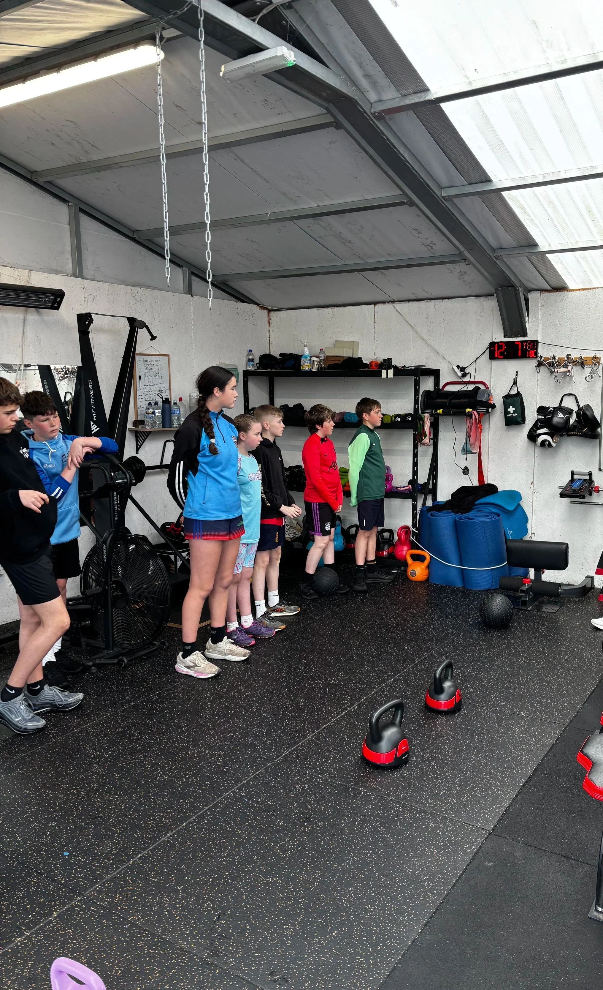 A group of young people standing in a line inside a gym, surrounded by fitness equipment like kettlebells, pulling machines, and mats, with a digital clock on the wall showing the time 12:79.