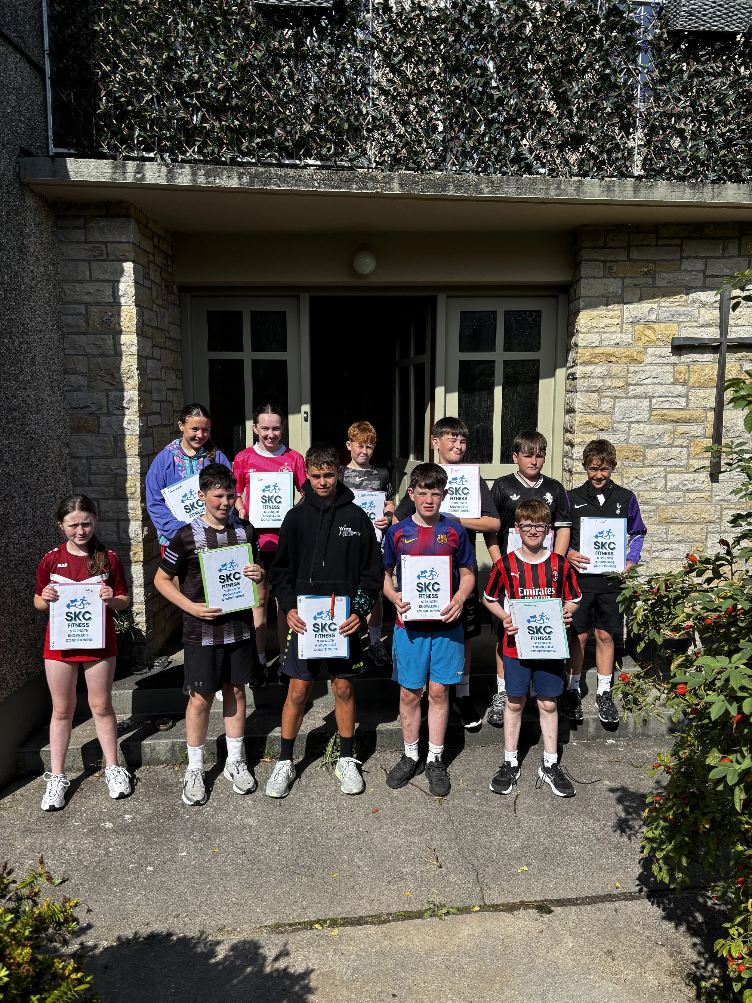 A group of children standing on the stairs in front of a house, some holding certificates that read 'SKC FITNESS,' with two girls at the back smiling, and boys in the front with serious faces. The children are wearing sports jerseys and athletic clothing.