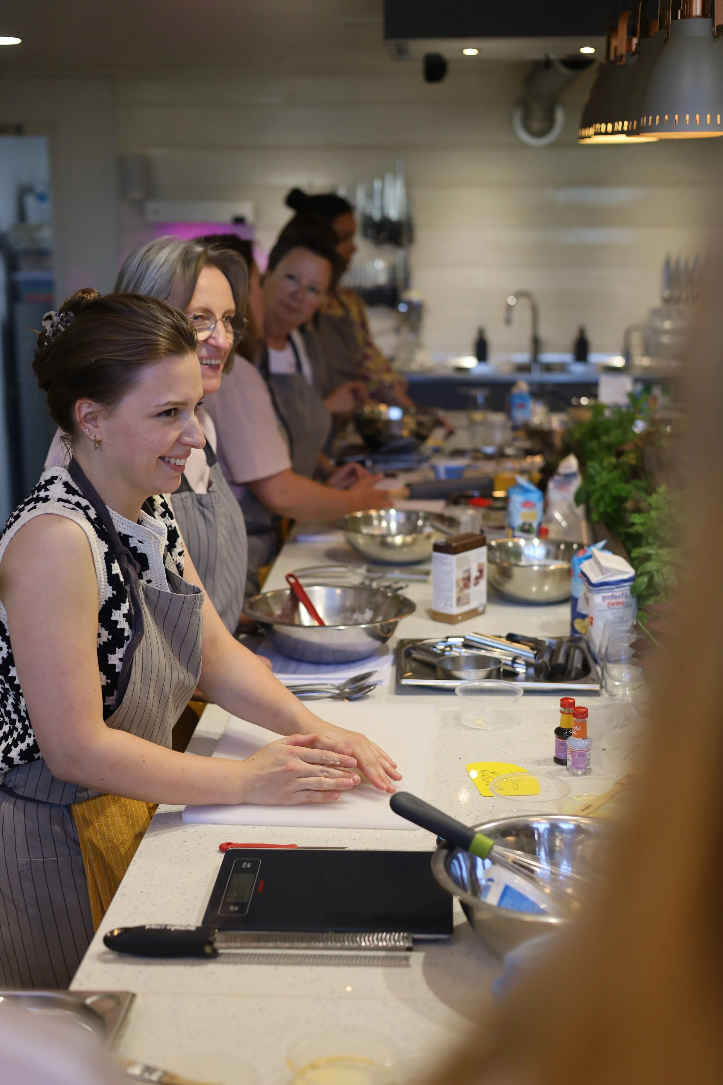 Een groep mensen die in een keuken aan het koken zijn, met de focus op een vrouw die haar handen wast op een aanrecht. In de omgeving staan bak- en bereidingsproducten, keukengerei en kruiden.