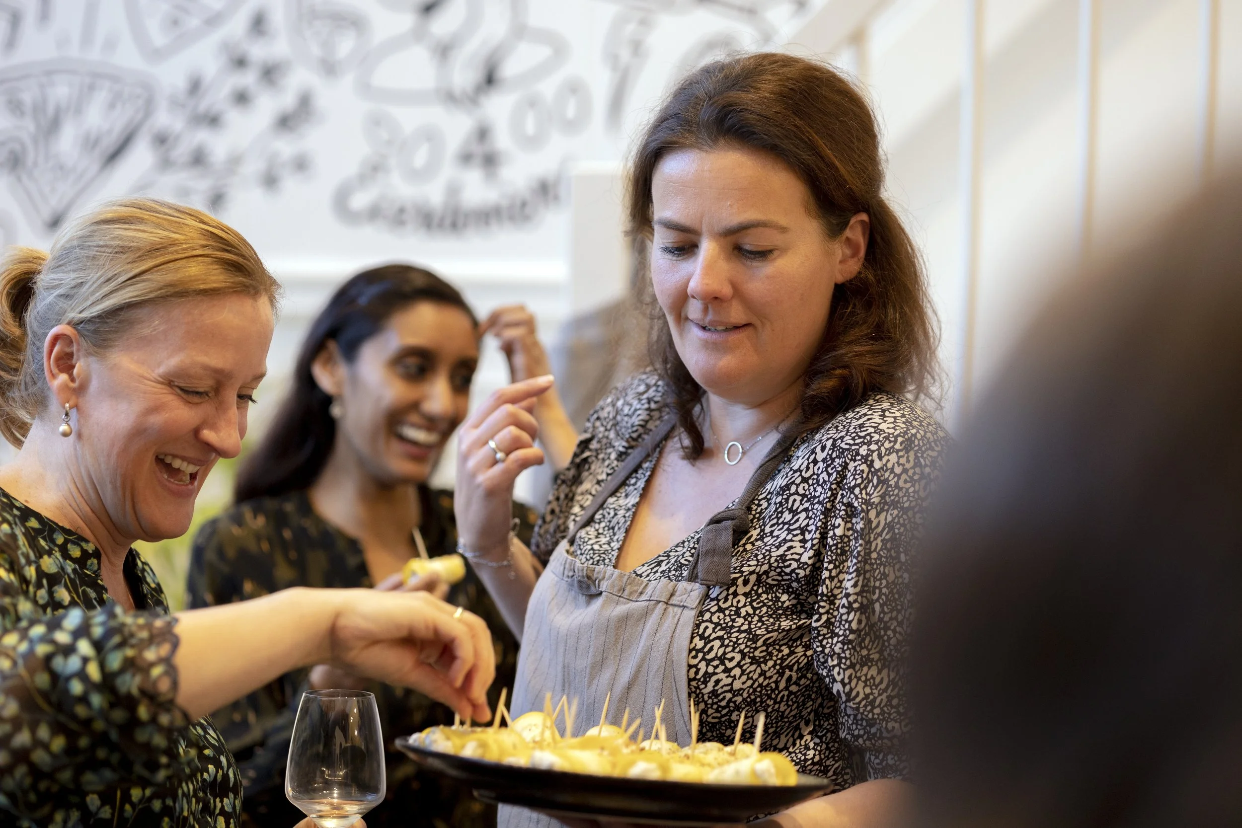 Vijf vrouwen die samen genieten van een etentje met hapjes en wijn, in een gezellige keuken of restaurant sfeer.