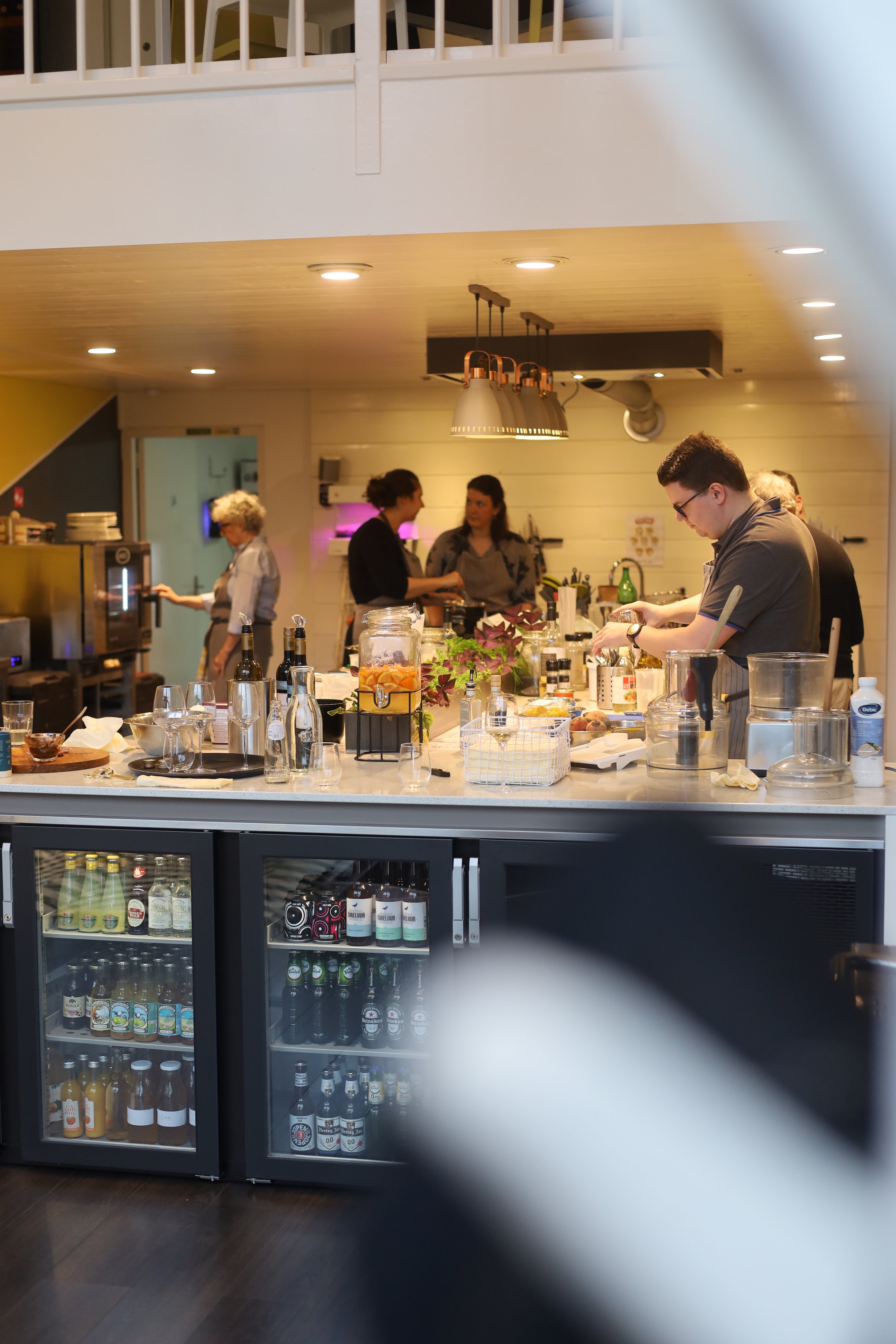 Binnen een restaurant of café met een bar, meerdere mensen werken achter de bar en bezig met het bereiden en serveren van drankjes. In de voorgrond staat een koelkast gevuld met diverse dranken.