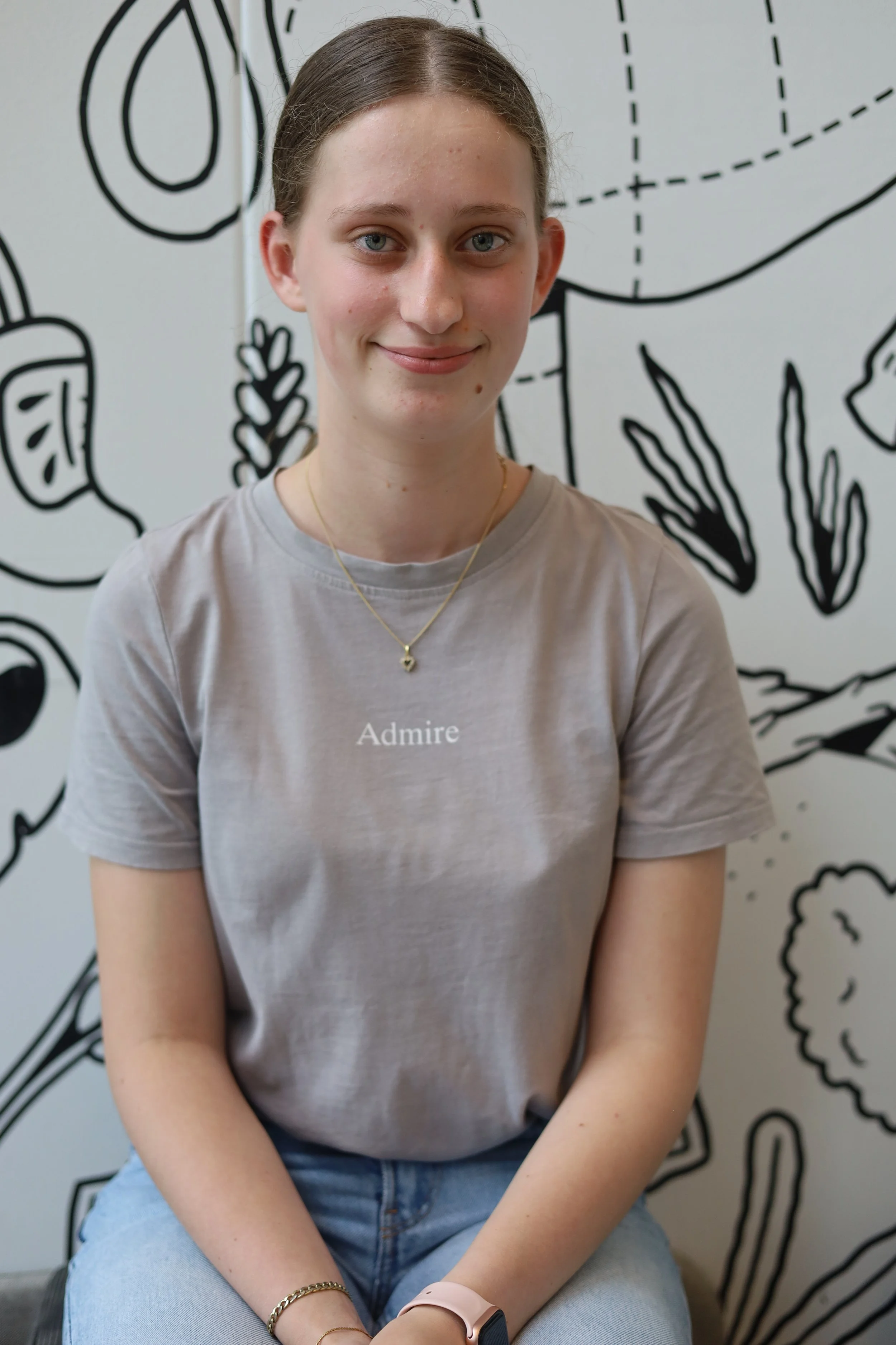 Een jonge vrouw met lichtbruin haar en een lichte huid zit voor een muur met zwart en wit tekeningen. Ze draagt een grijs T-shirt met de tekst 'Admire', een gouden ketting, een gouden armband en een smartwatch.