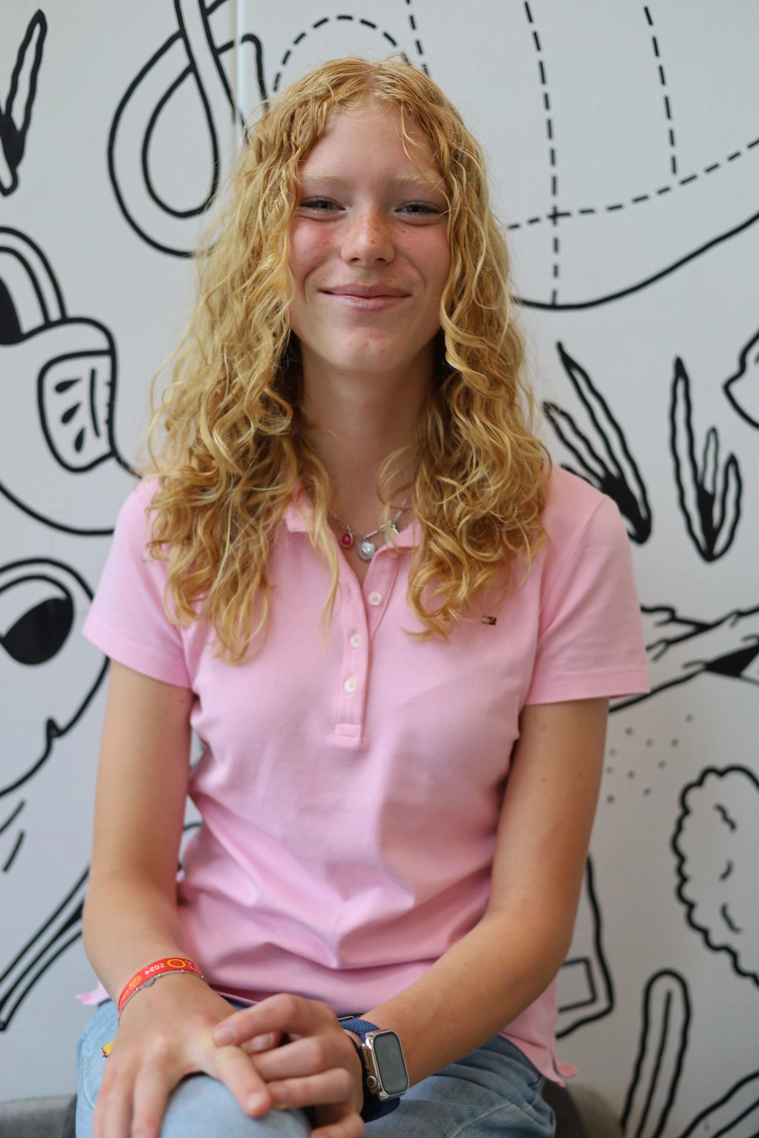 Een lachende jonge vrouw met gekruld rood haar zit voor een muur met zwart-witte schetsachtige tekening van bloemen en planten, draagt een roze polo en een smartwatch.