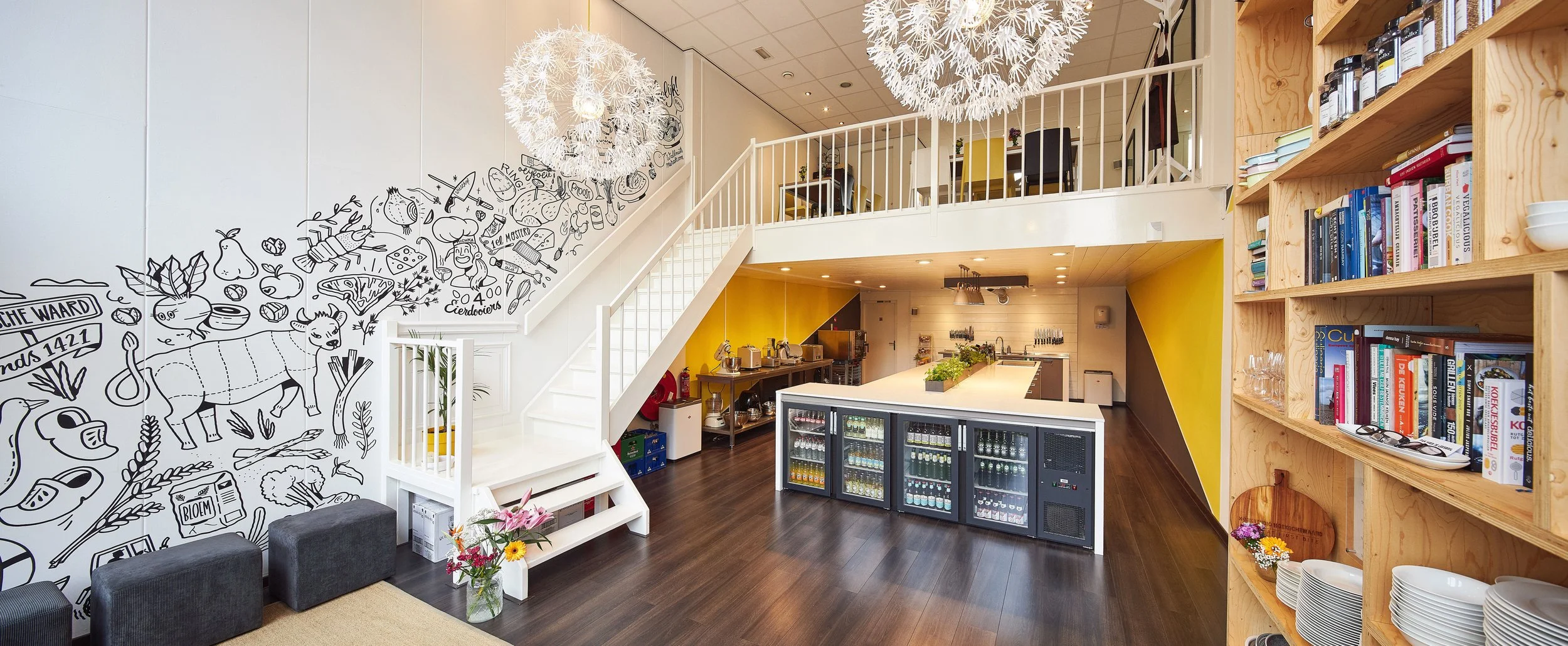 Interieur van een modern café of restaurant met ronde witte hanglampen, een boekenkast gevuld met boeken, een bar met koelkasten, en een kleurrijke muur met tekeningen en tekst.