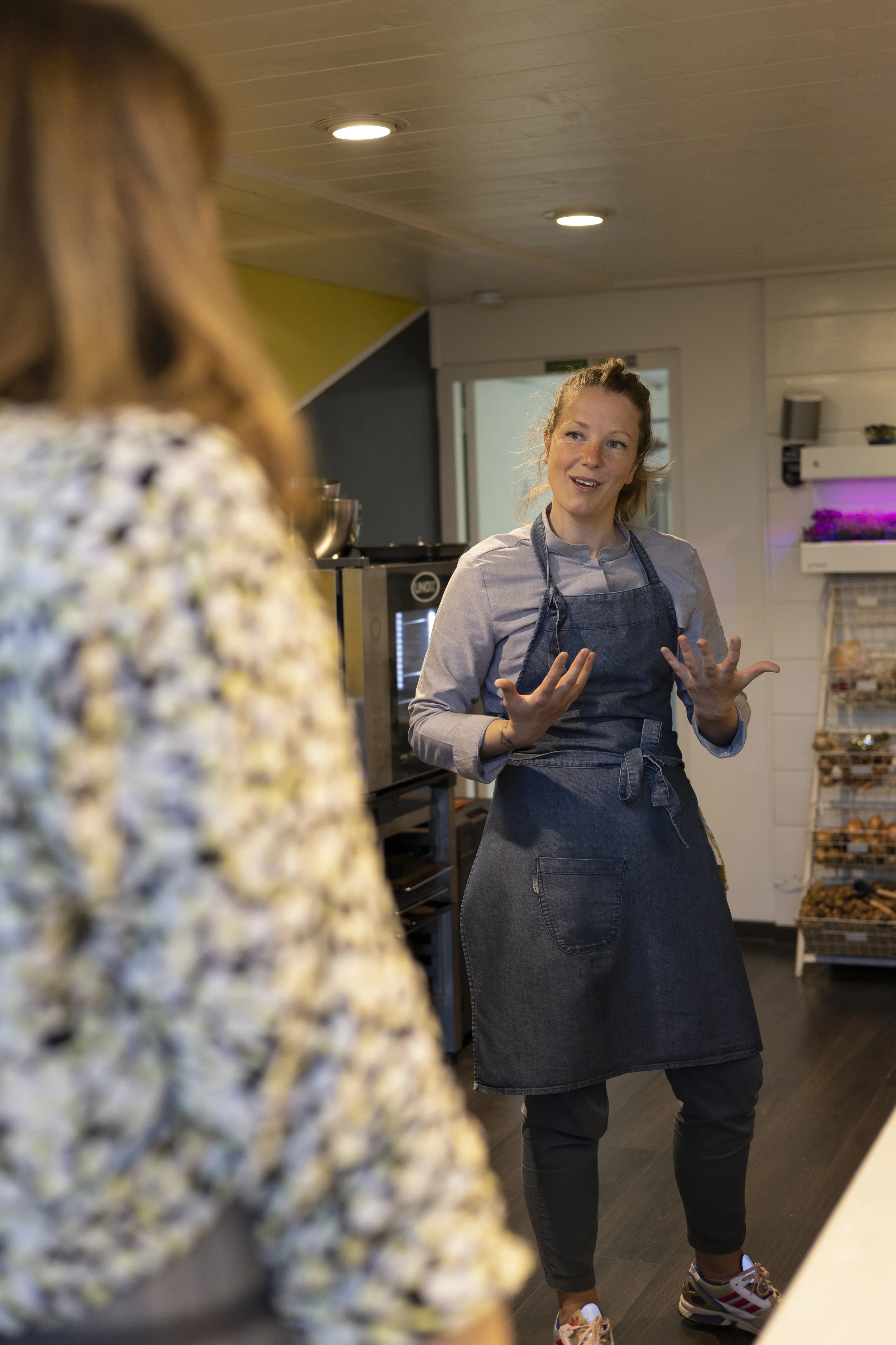 Vrouw die uitlegt in een keuken of winkel, met oven en planten in de achtergrond.