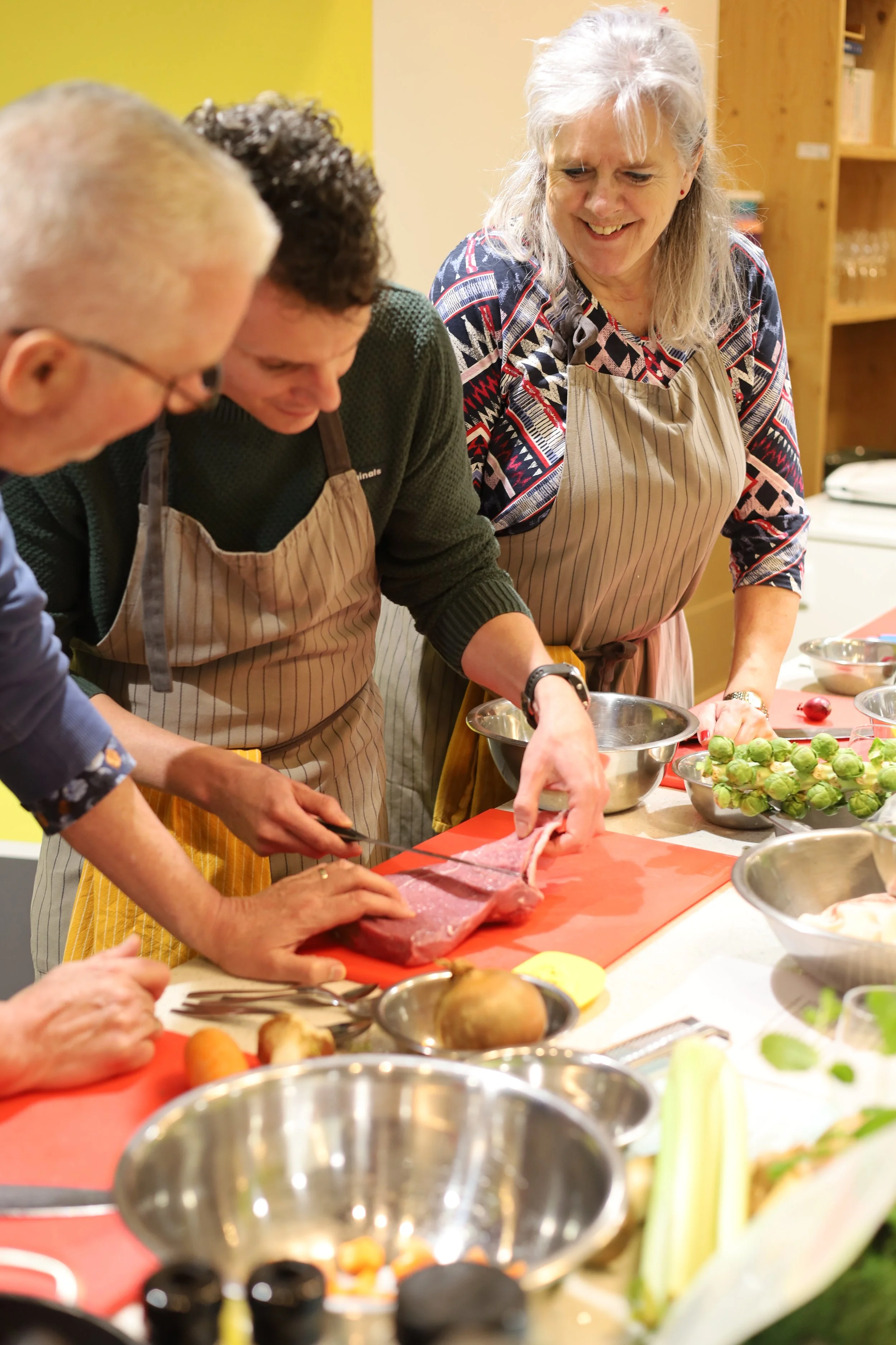 Mensen die gezamenlijk koken in een keuken, met groenten, vlees en keukenbenodigdheden op het aanrecht.