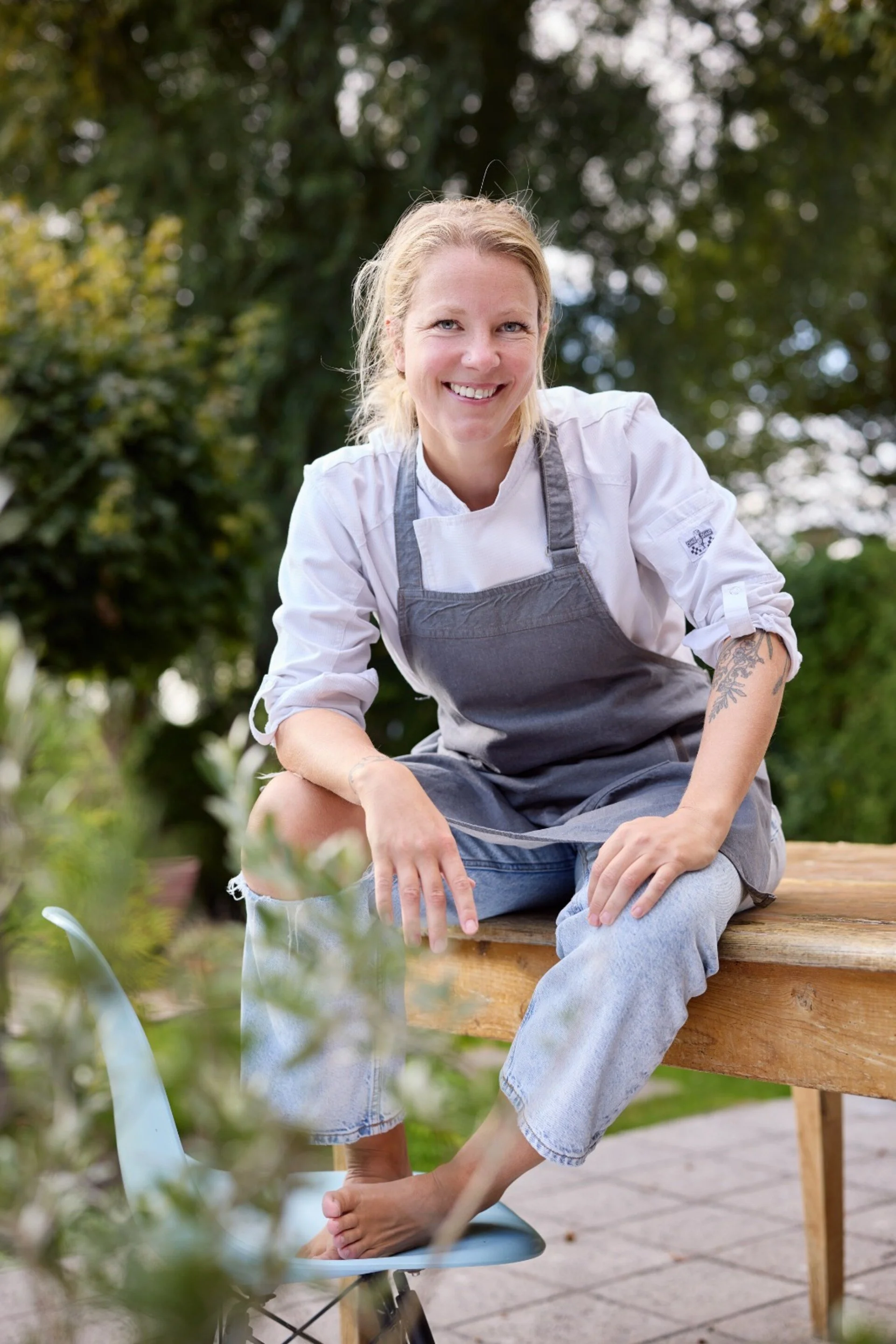 Een vrouw met blond haar in een witte blouse en een grijze schort, zittend op een houten bank buiten, lachend naar de camera, omringd door groen blad en bomen.