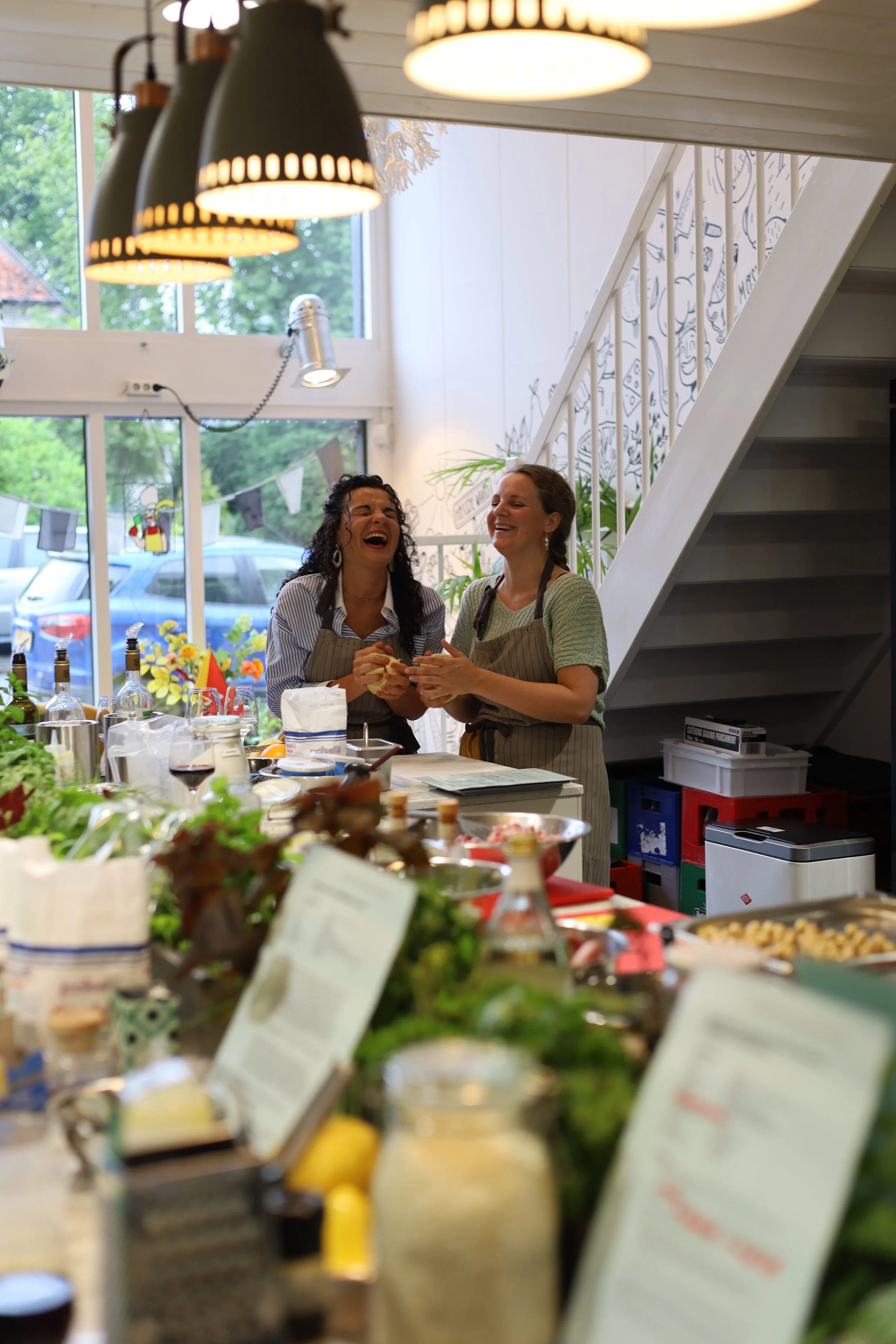 Twee vrouwen lachen en praten in een keuken, omgeven door gerechten en ingrediënten.