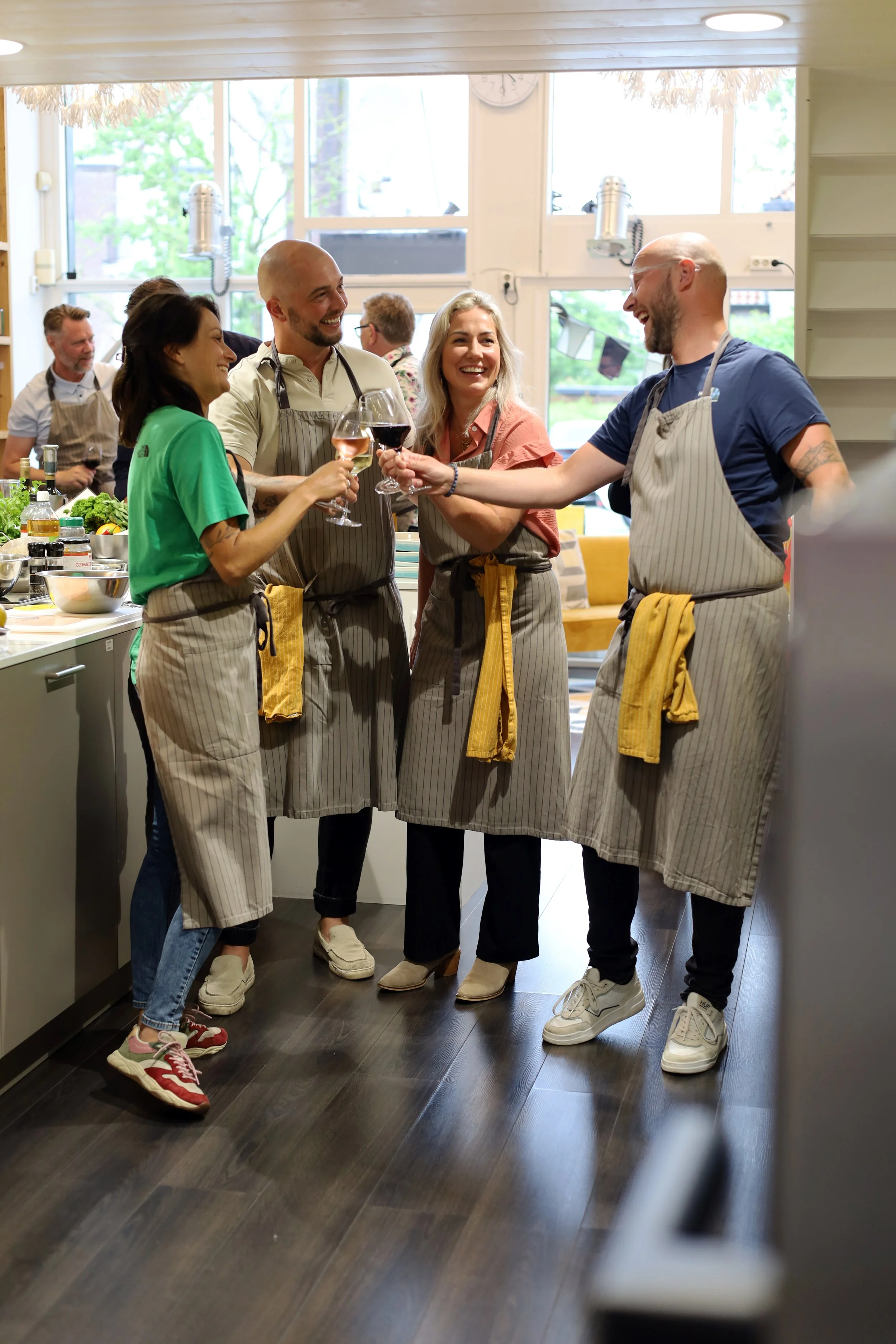 Vijf mensen in een keuken, allemaal dragen ze schorten. Ze lachen en proosten met wijnglazen, er lijkt een gezellige sfeer te zijn.
