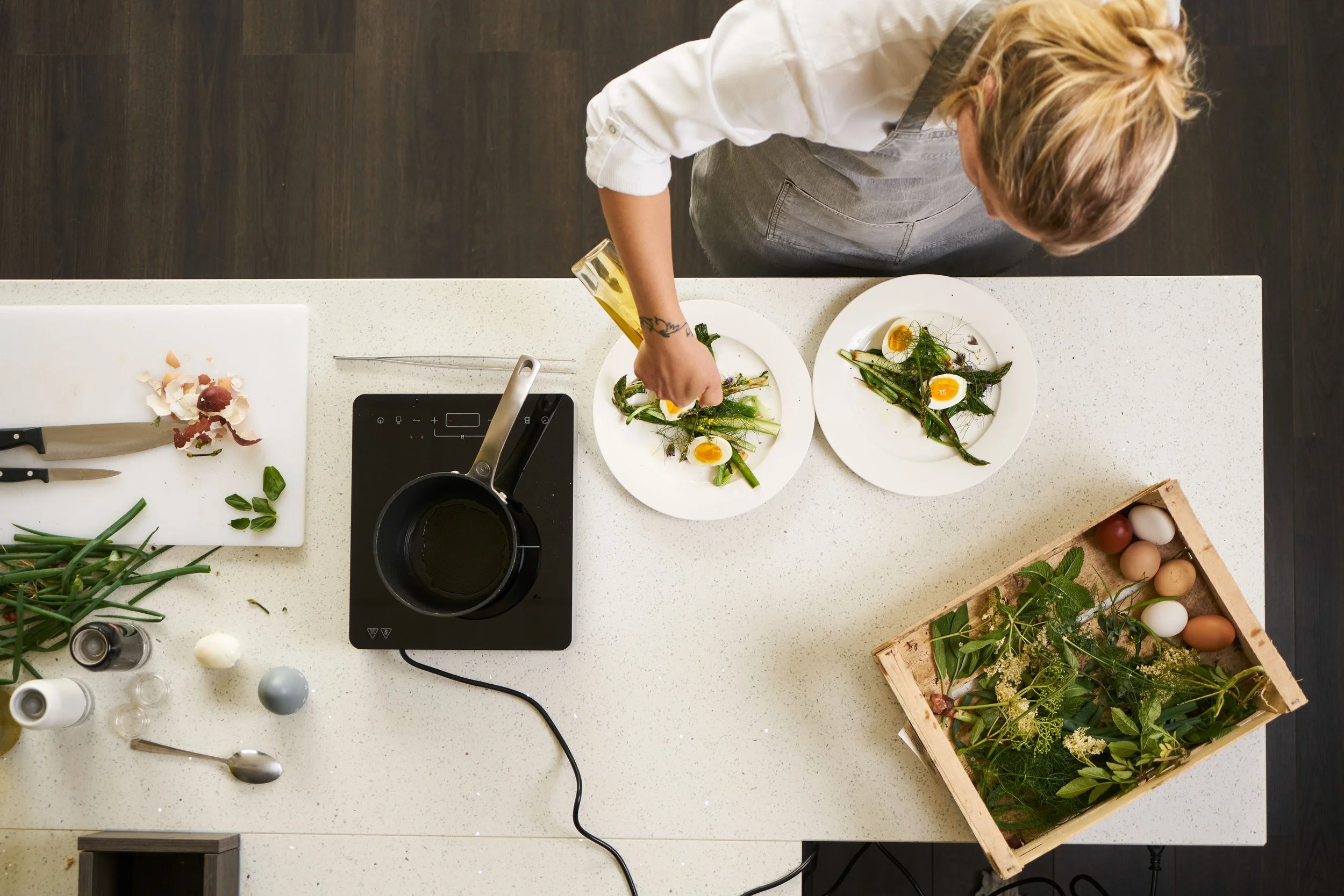 Bovenaanzicht van een persoon die een salade bereidt op een witte keukentafel, met diverse ingrediënten en keukengerei eromheen.
