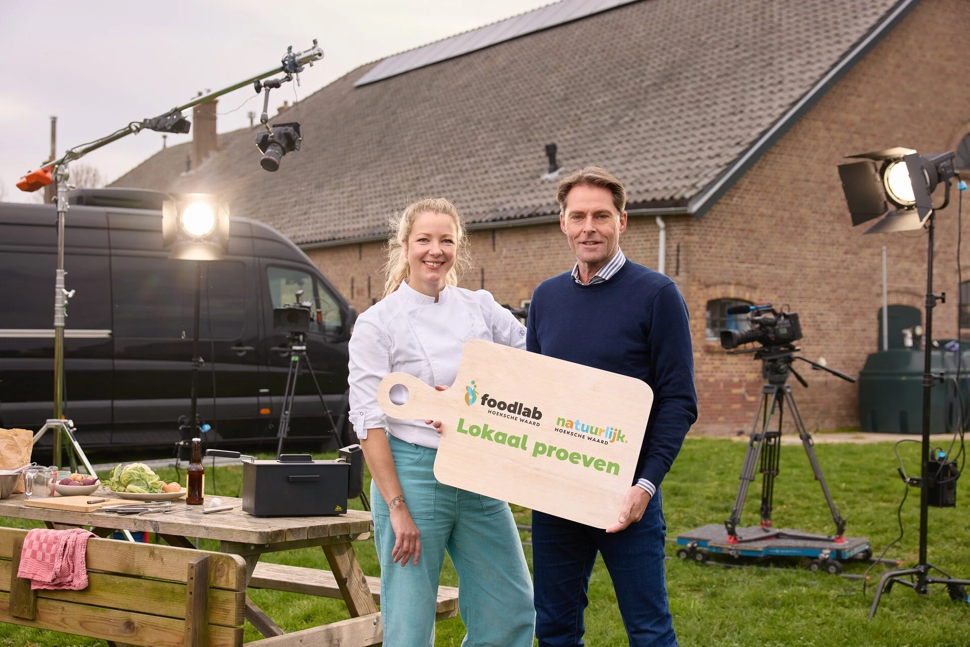 Twee mensen, een vrouw en een man, staan buiten voor een huis met zonnepanelen op het dak. Ze houden een grote houten tekenbord vast met de tekst 'Lokale proef' en logo's van Foodlab en Natuurlijke. Op de achtergrond is film- en fotografiemateriaal te zien, zoals studioverlichting, camera's en statieven.