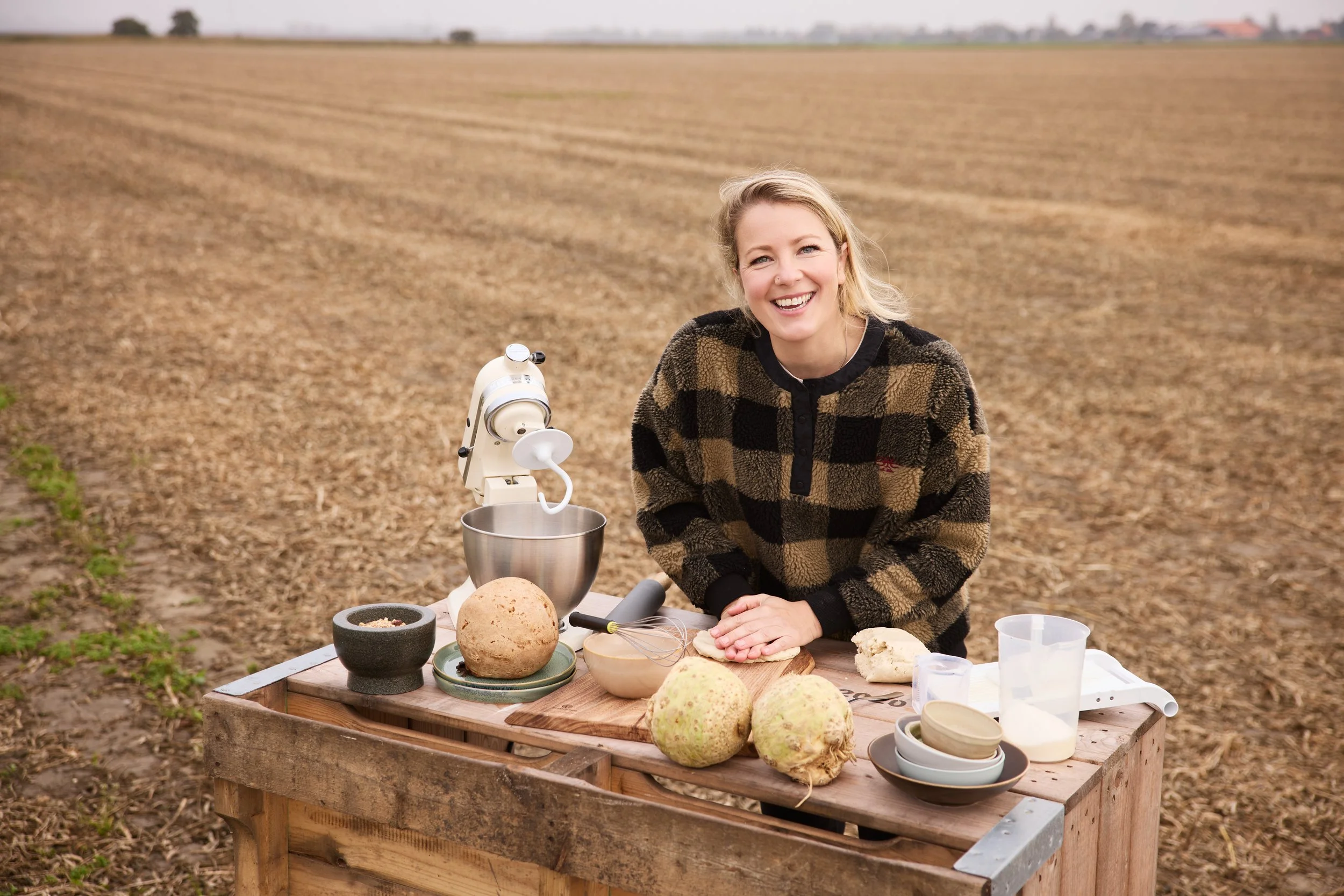 Vrouw die buiten op een tafel met bakproducten en keukengerei staat, met een akkerland op de achtergrond.