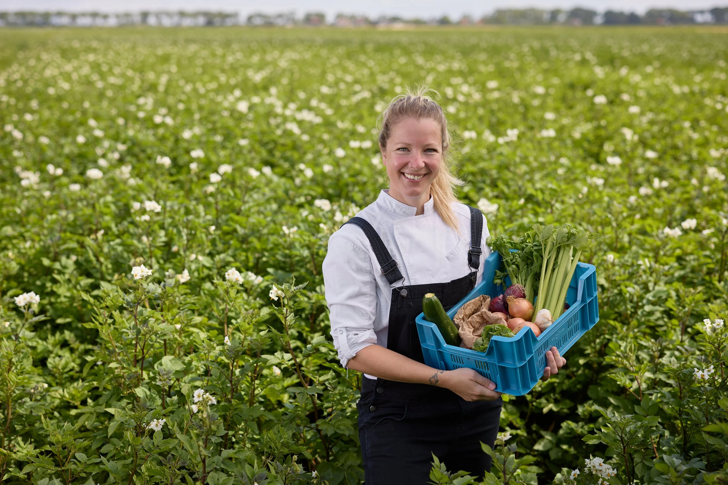 Vrouw in een veld met witte bloemen, lachend en een blauwe krat met verse groenten zoals uien, komkommer, selderij, knoflook en aardappelen vasthoudend.