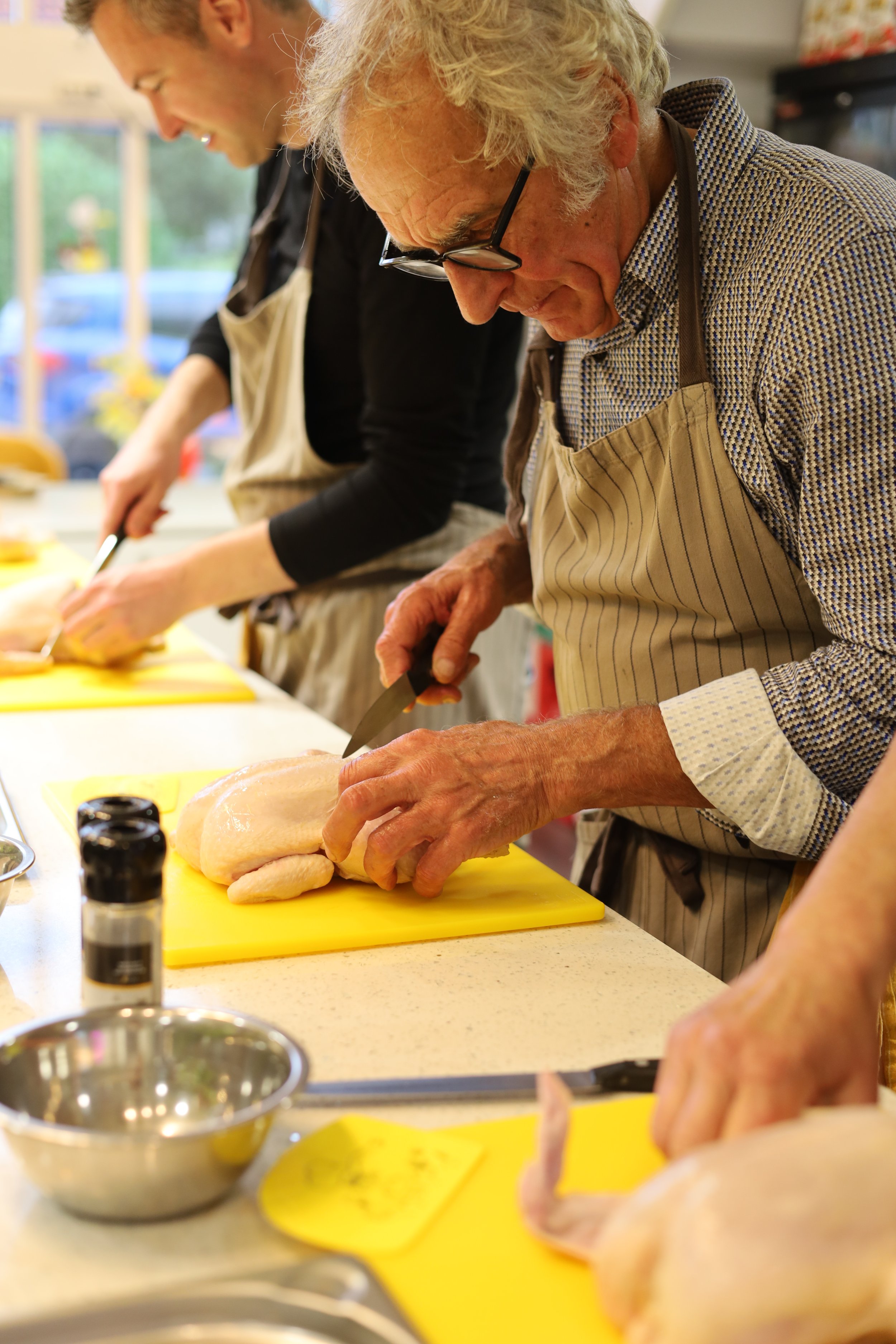 Twee oudere mannen bereiden kipfilets voor in een keuken, een met een kruidenschaal en de ander met een snijplank en mes.