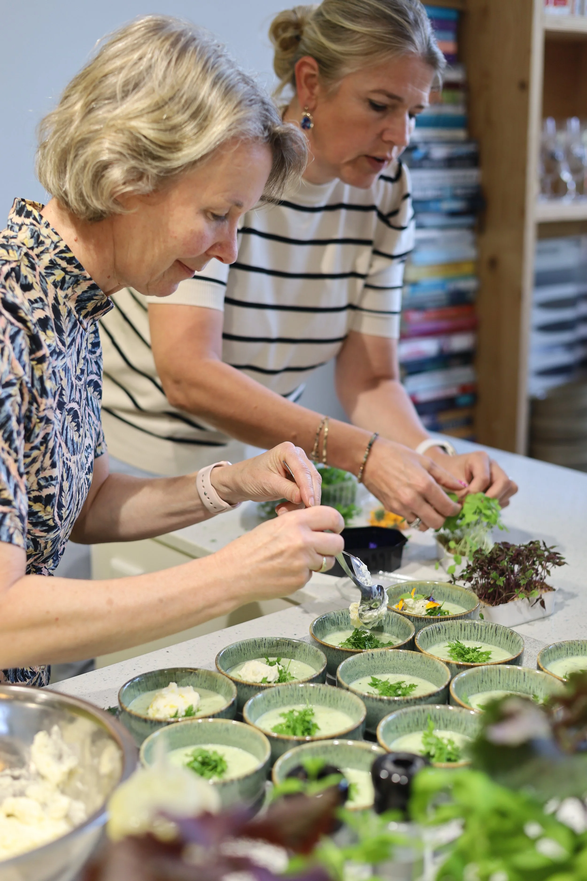 Twee vrouwen maken gerechten met kruiden en salades op een keukenblad, met verschillende kleine bowls gevuld met ingrediënten.