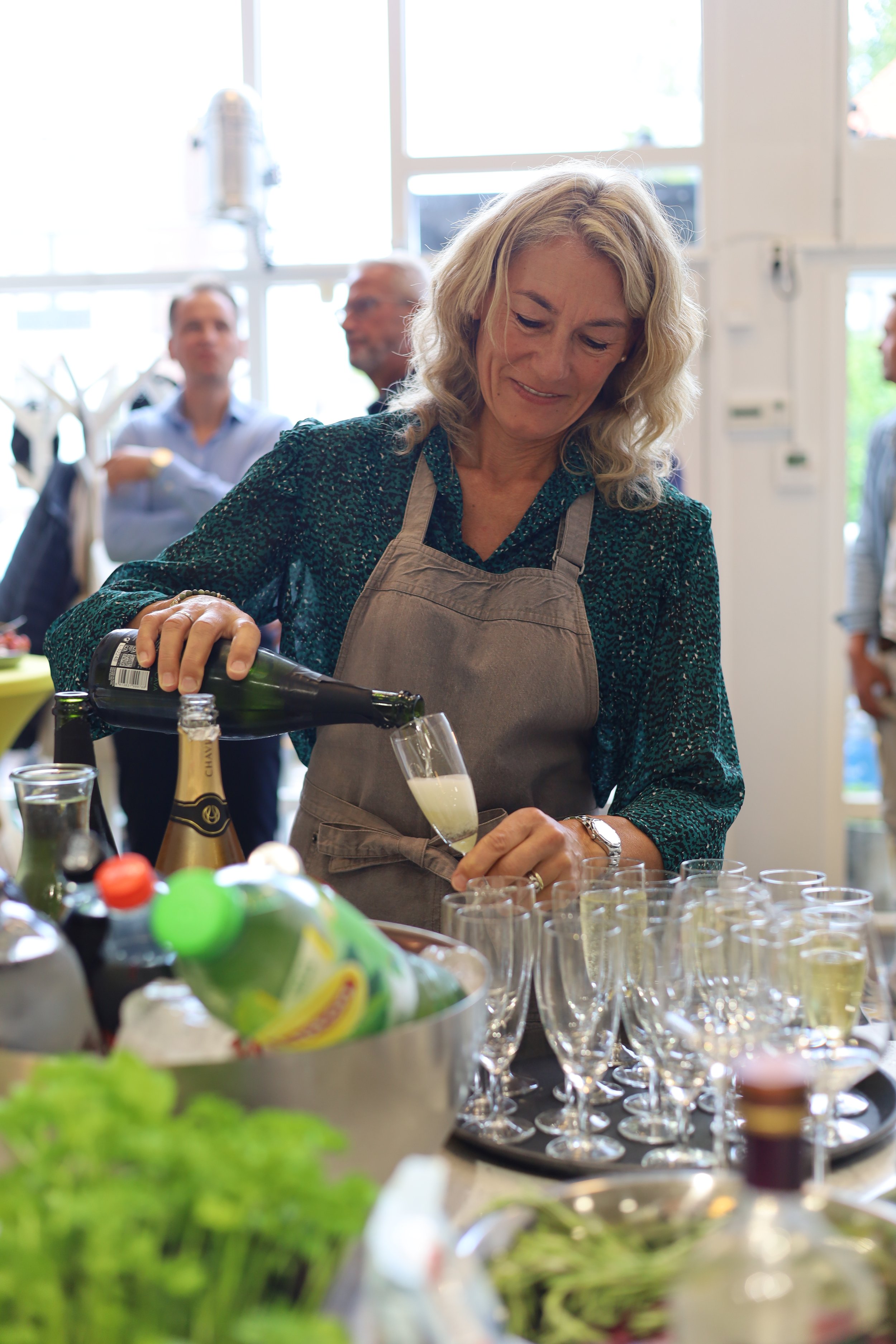 Een vrouw schenkt een drankje in een glazen tijdens een sociale bijeenkomst, met vier andere mensen op de achtergrond in een lichte kamer met grote ramen.