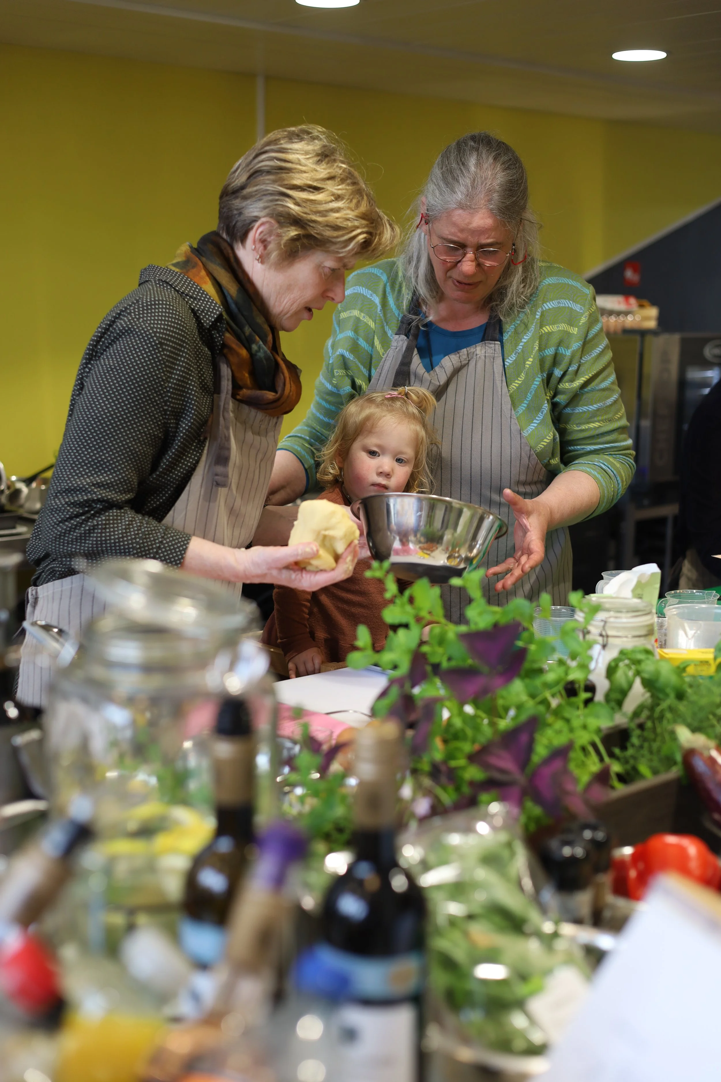 Twee oudere vrouwen en een klein meisje die samen in een keuken bereiden, met diverse groenten en keukengerei op de tafel.