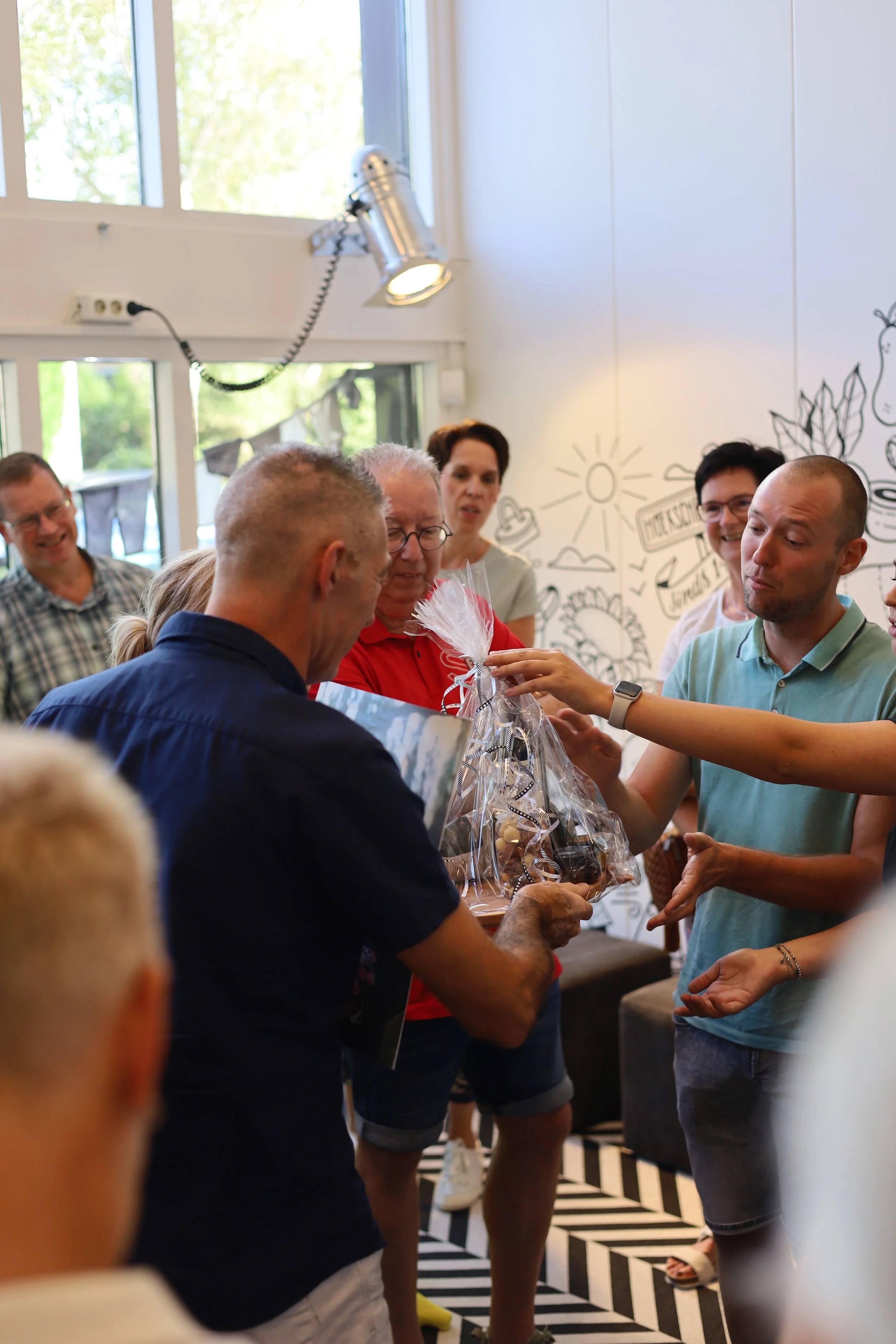 Een groep mensen die samen een cadeau uitpakken in een interieur met muurschilderingen en grote ramen.