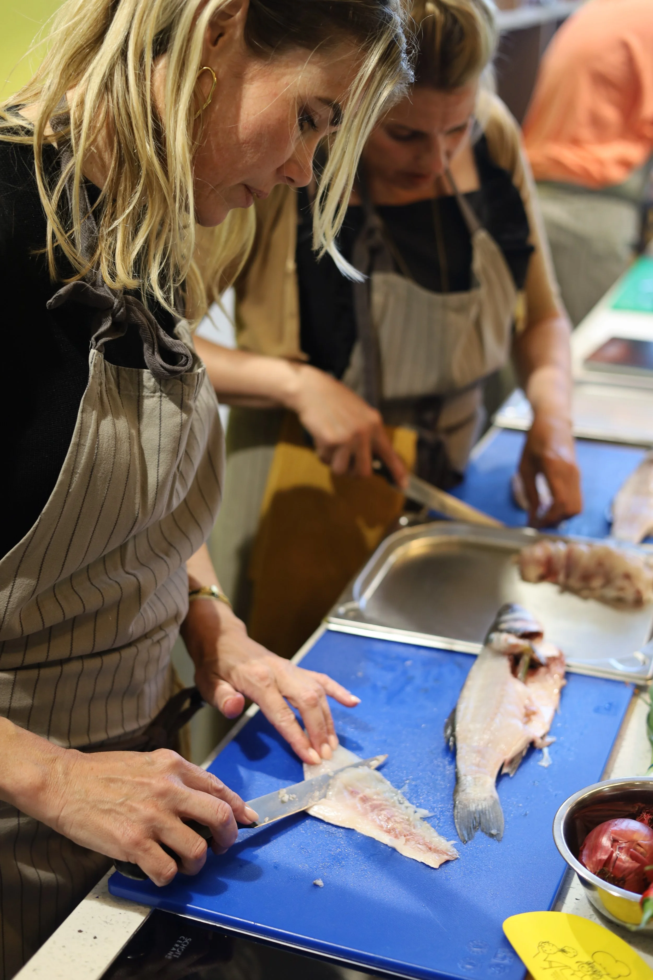 Vrouw snijdt vis op een snijplank in een keuken, terwijl een andere vrouw naast haar voorbereidt.