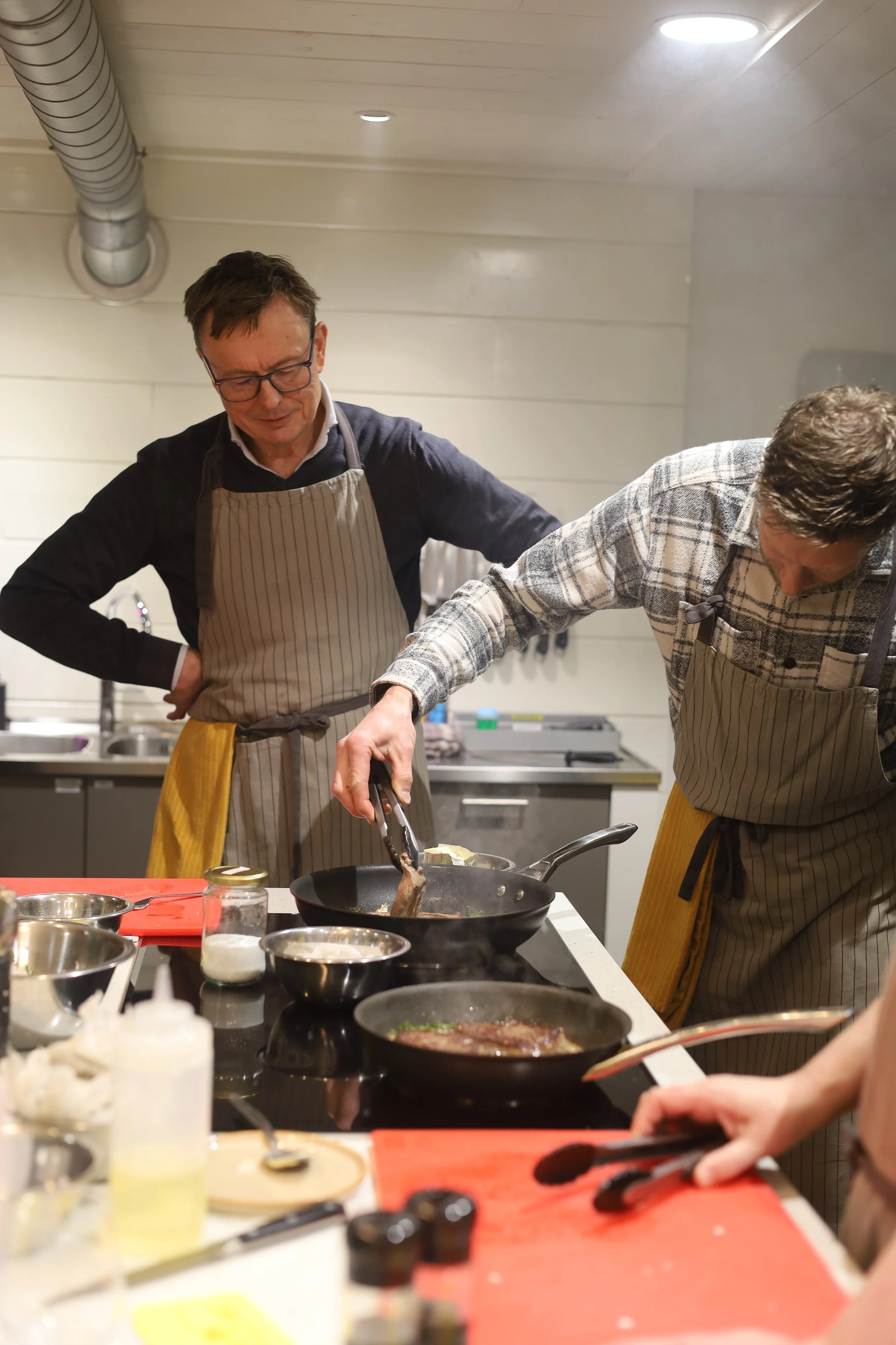 Twee mannen koken samen in een keuken. Ze dragen aprons en zijn bezig met het bereiden van eten in pannen.