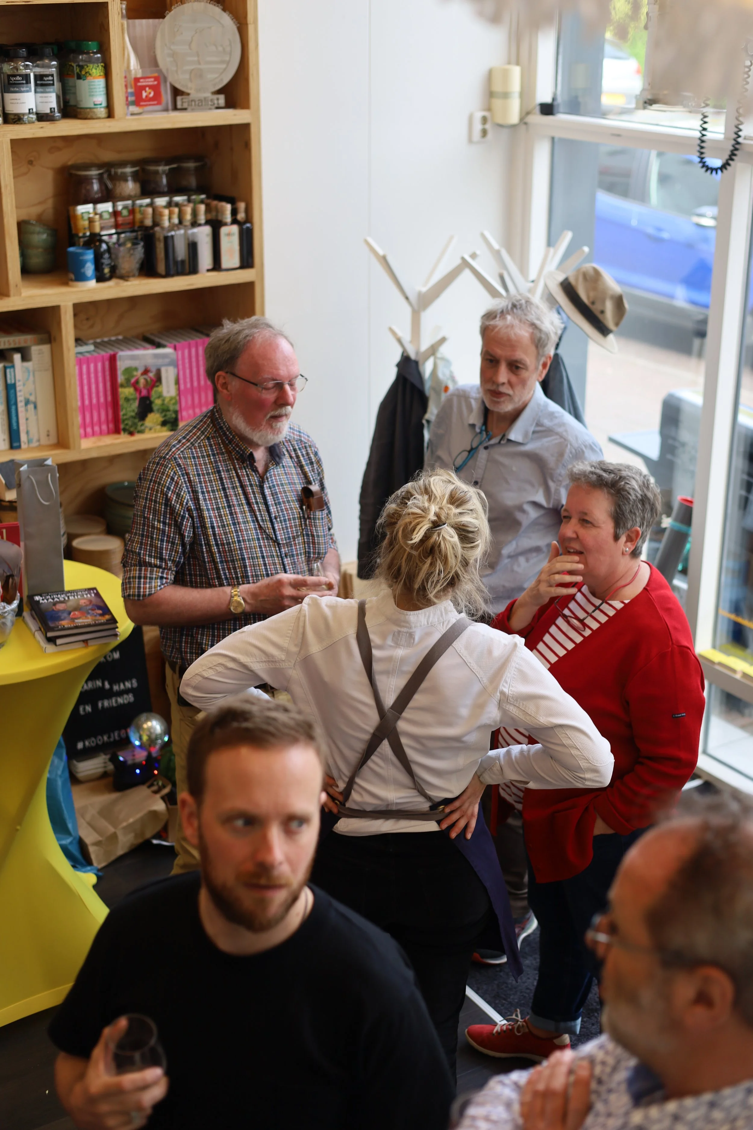 Groep mensen die in een winkel praten, met planken met kruiden en boeken op de achtergrond, en een grote raampartij met uitzicht op de straat.