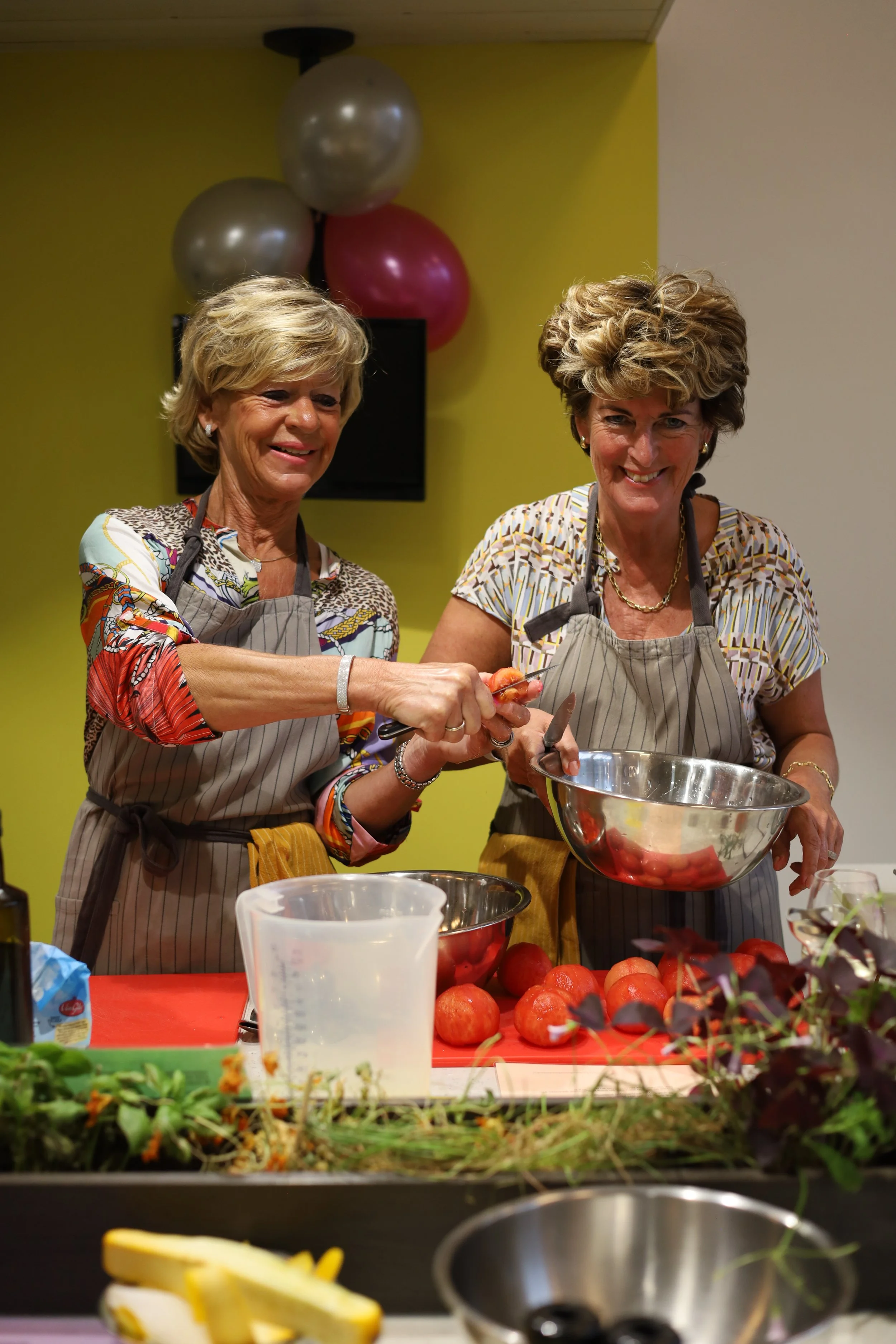 Twee vrouwen koken samen in een keuken, ze dragen schorten en lijken plezier te hebben, met balloon decoraties op de achtergrond.